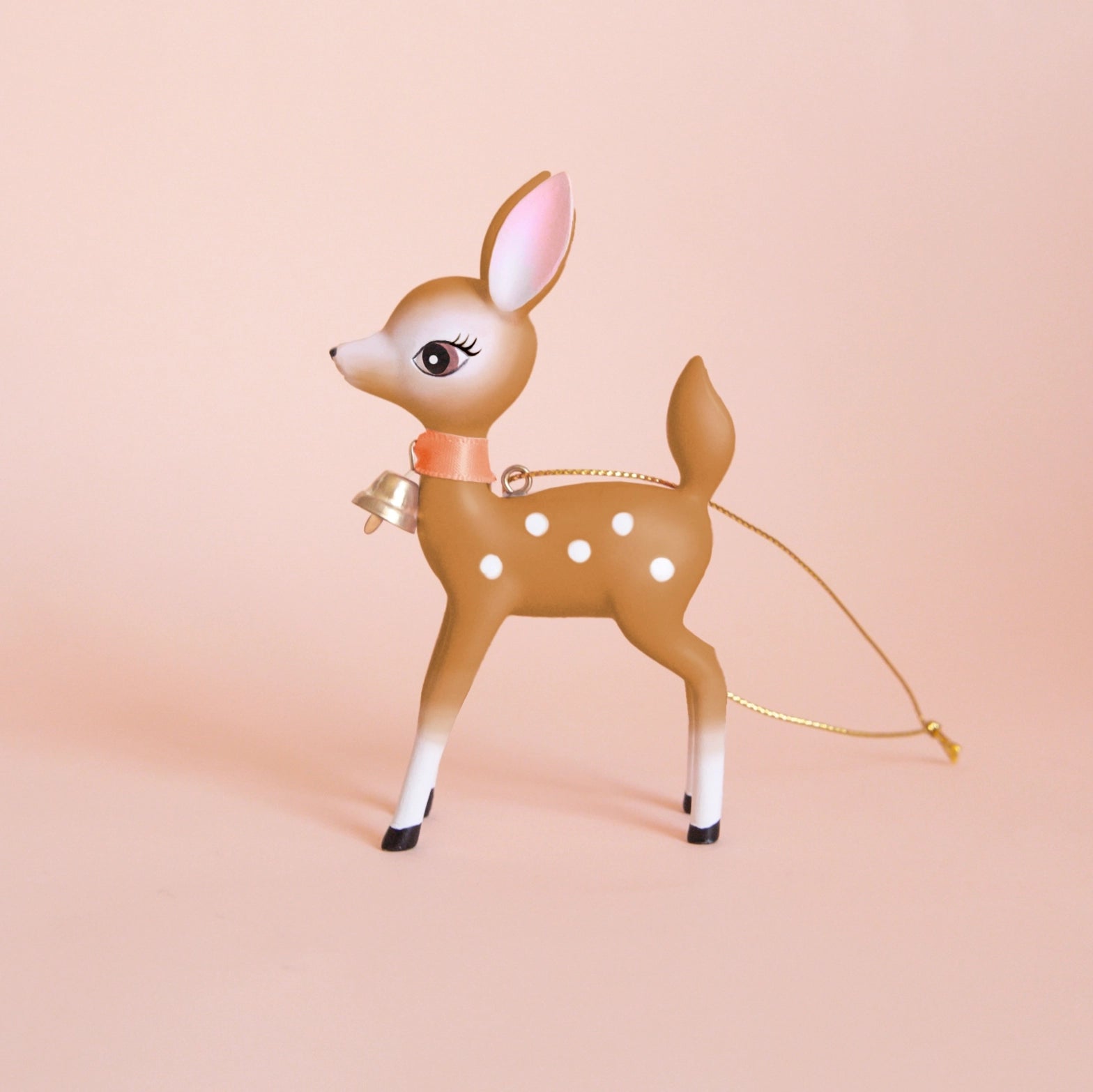 On a peachy background is a brown retro deer ornament with white details and spots and a gold bell on the front of its neck along with a gold string loop for hanging.