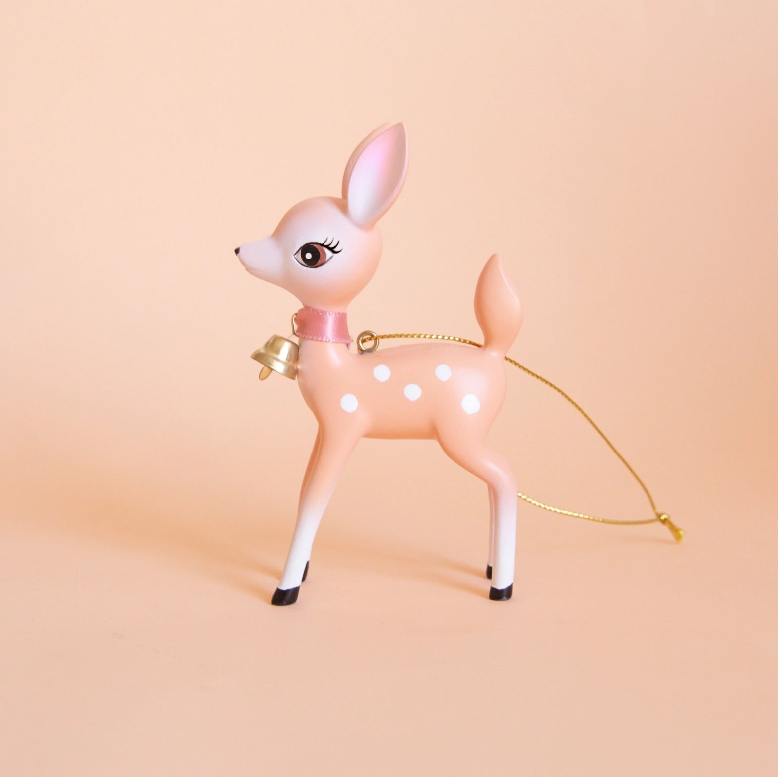 On a peach background is a peachy pink retro deer ornament with white details and spots and a gold bell on the front of its neck along with a gold string loop for hanging.