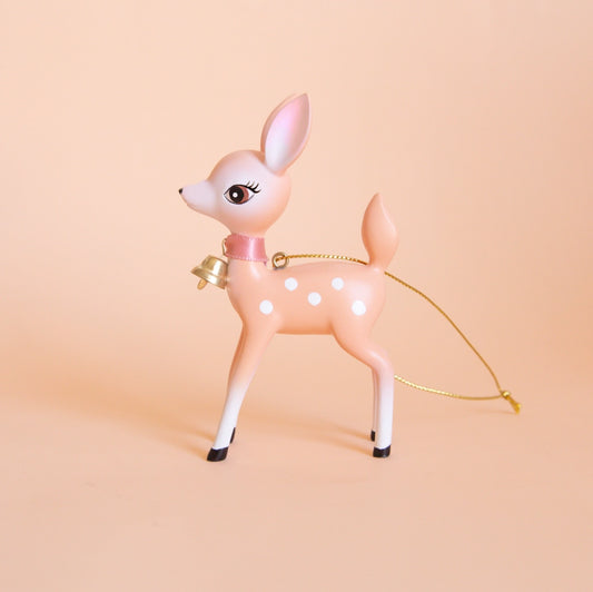 On a peach background is a peachy pink retro deer ornament with white details and spots and a gold bell on the front of its neck along with a gold string loop for hanging.