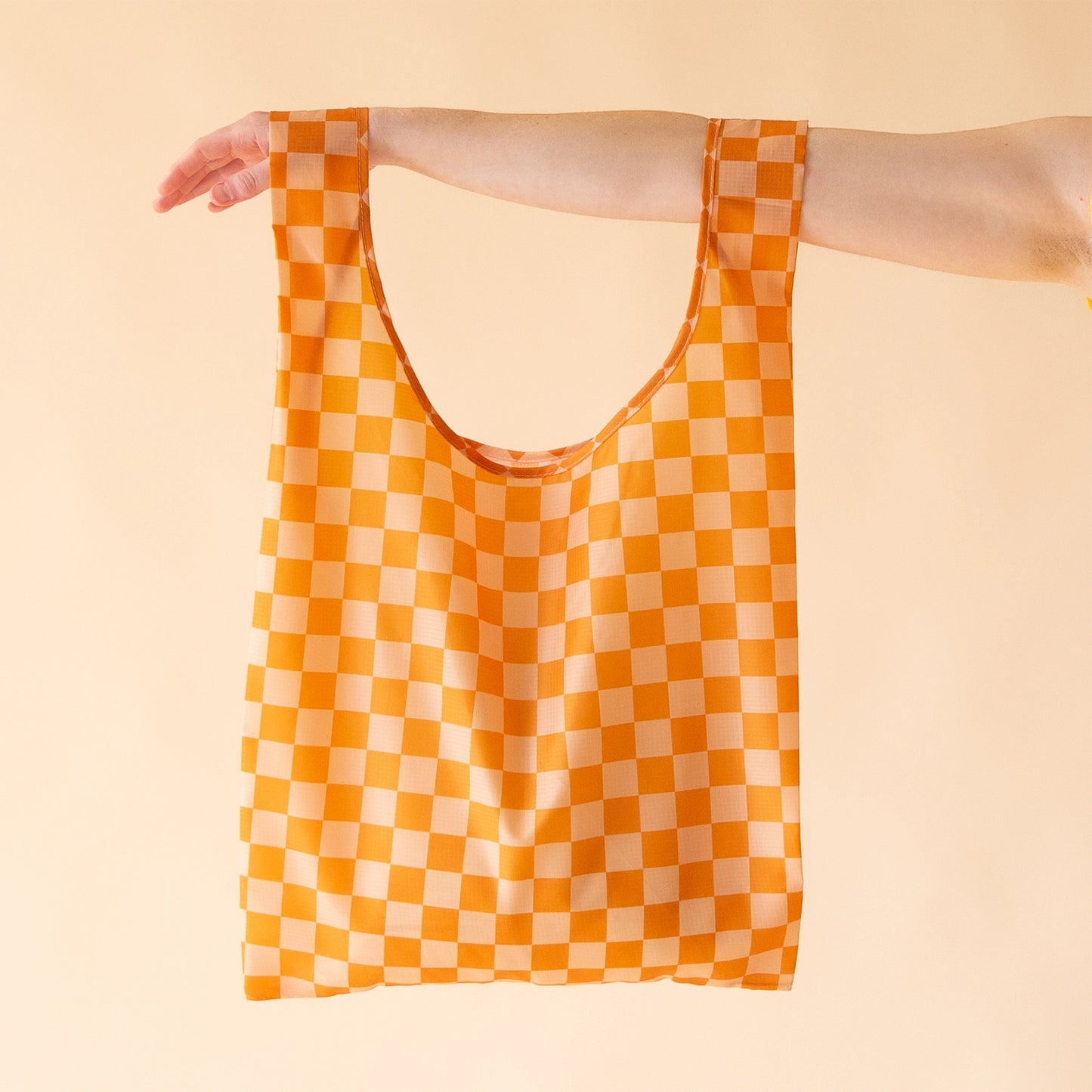 On a light peach background is an orange checkered nylon tote bag being held by a models arm.