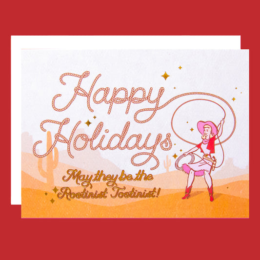 Cowgirl swinging a lasso with rope style font that reads "Happy Holidays May they be the rootinist tootinist!" There is a cactus and mountain range background behind her.  Card is laying on a pink background paper.