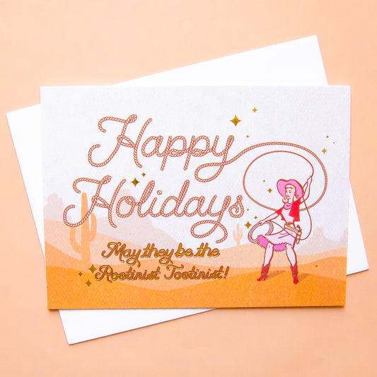 Cowgirl swinging a lasso with rope style font that reads "Happy Holidays May they be the rootinist tootinist!" There is a cactus and mountain range background behind her.  Card is laying on a pink background paper.