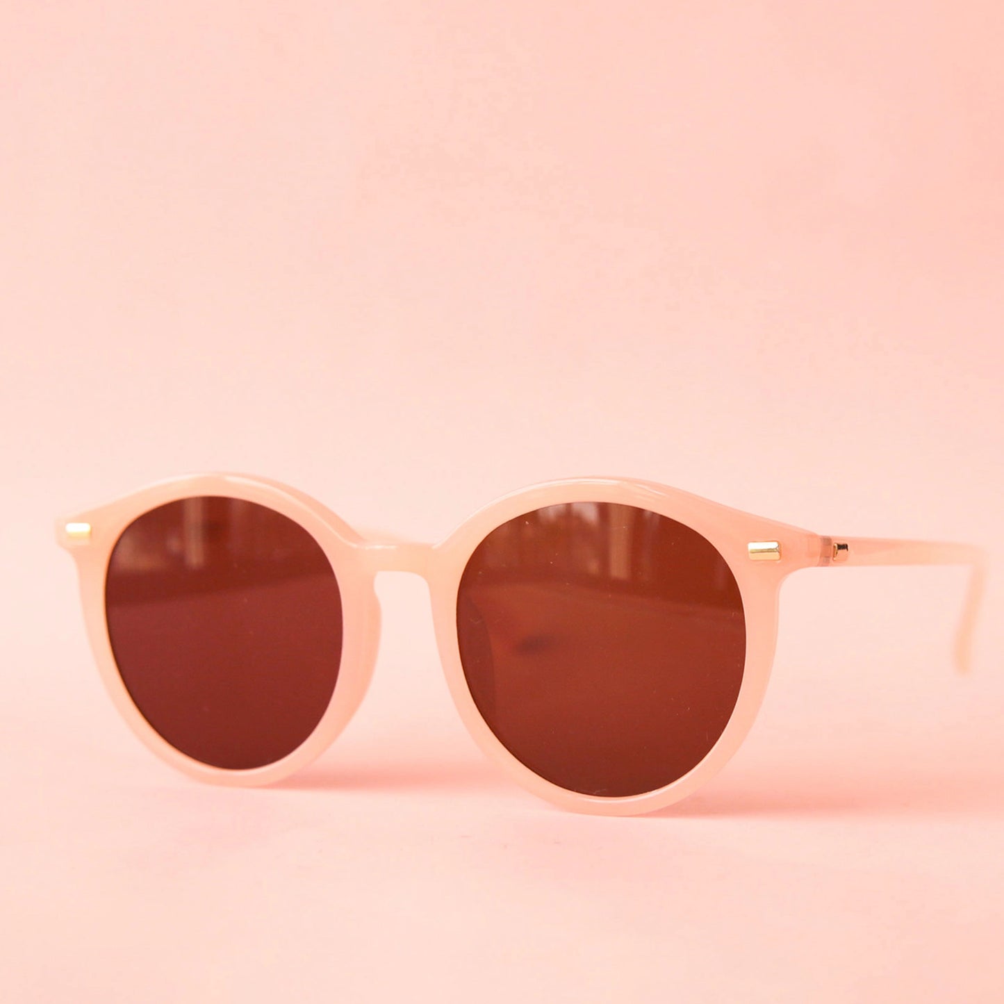 On a pink backgrounds a pink framed pair of round sunglasses with brown lenses. 