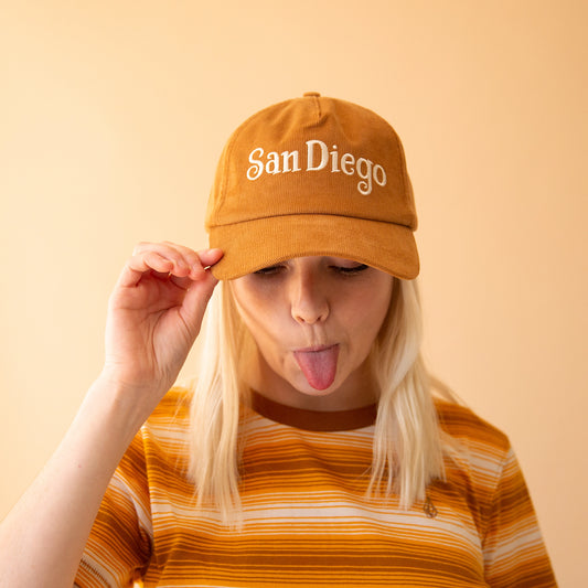 On a light peach background is a model wearing a toffee colored corduroy baseball hat with white stitching that reads, "San Diego".