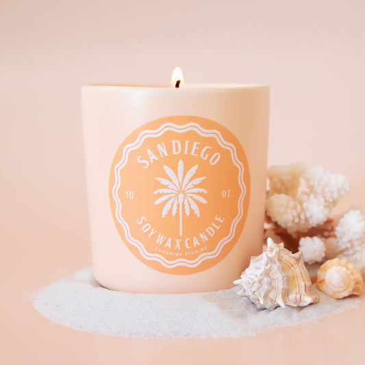 A light tan ceramic candle jar with a light orange circle label in the center with a palm tree graphic and text that reads, 'San Diego Soy Wax Candle'. 