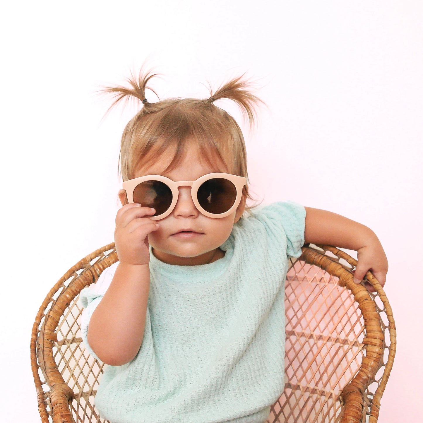 On a white background is a children's model wearing a light pink pair of round sunglasses. 