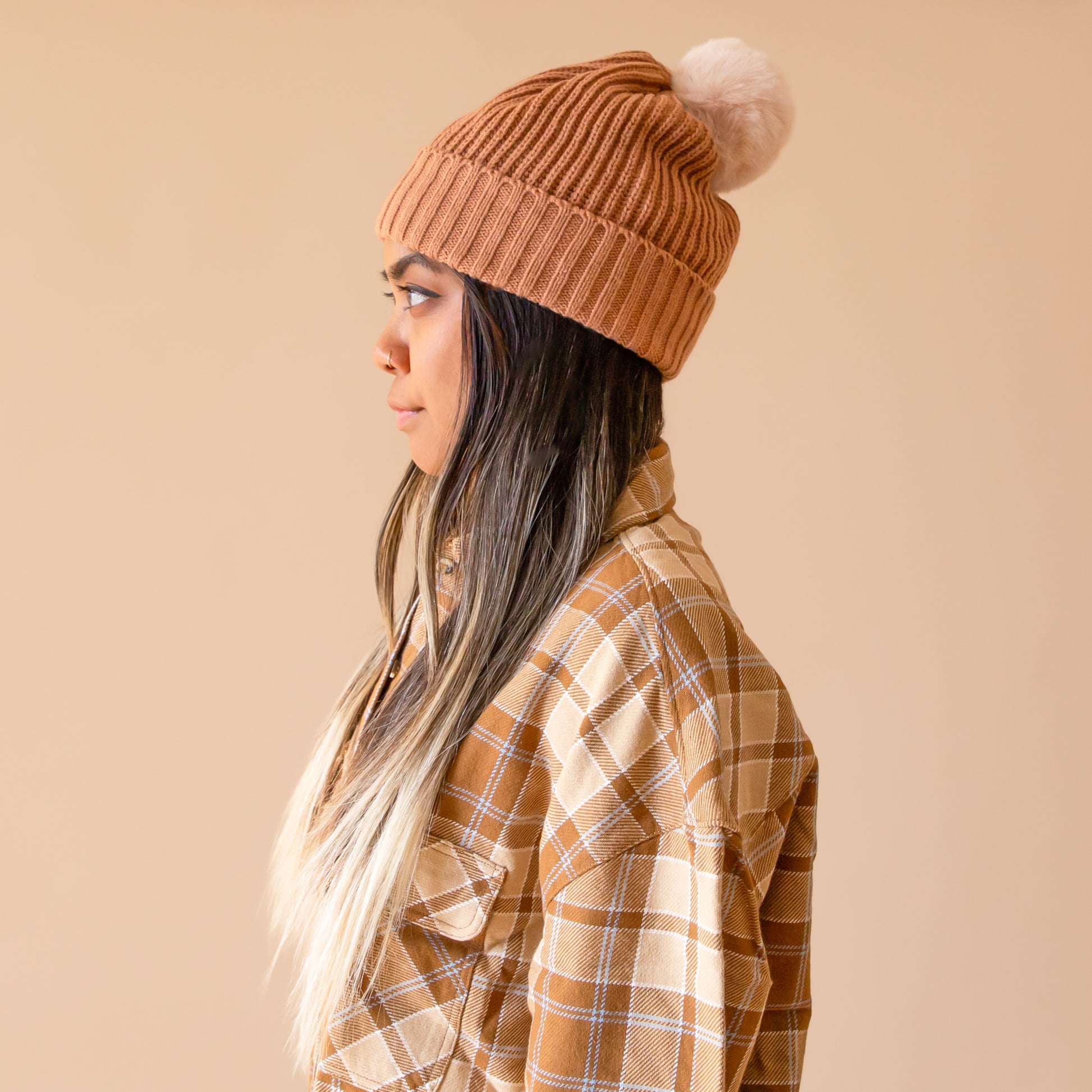 A warm brown colored beanie with a soft pom pom on top worn by a model with long brunette hair wearing a flannel shirt.