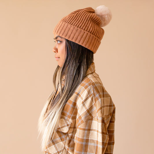 A warm brown colored beanie with a soft pom pom on top worn by a model with long brunette hair wearing a flannel shirt.