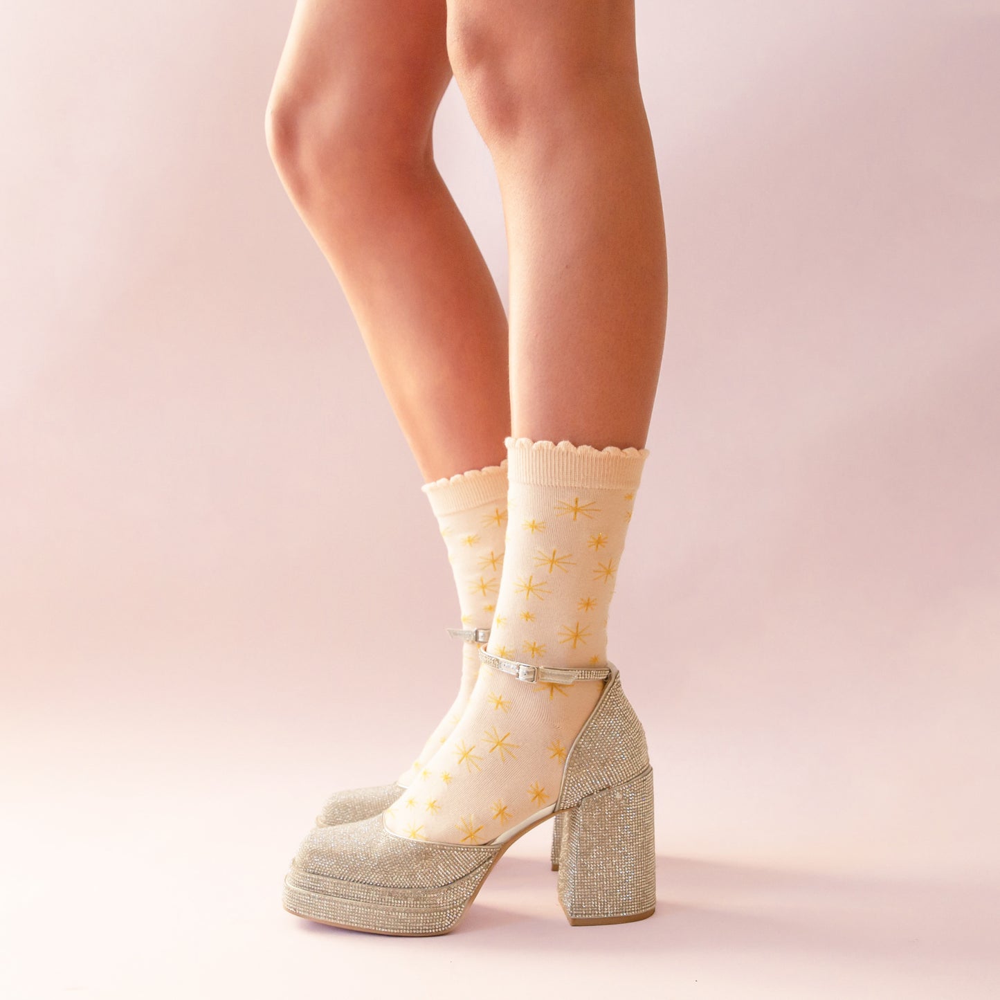 Close-up of crossed legs wearing sheer cream socks with yellow starburst prints and ruffled cuffs, paired with glittery silver platform Mary Jane heels against a soft pink background