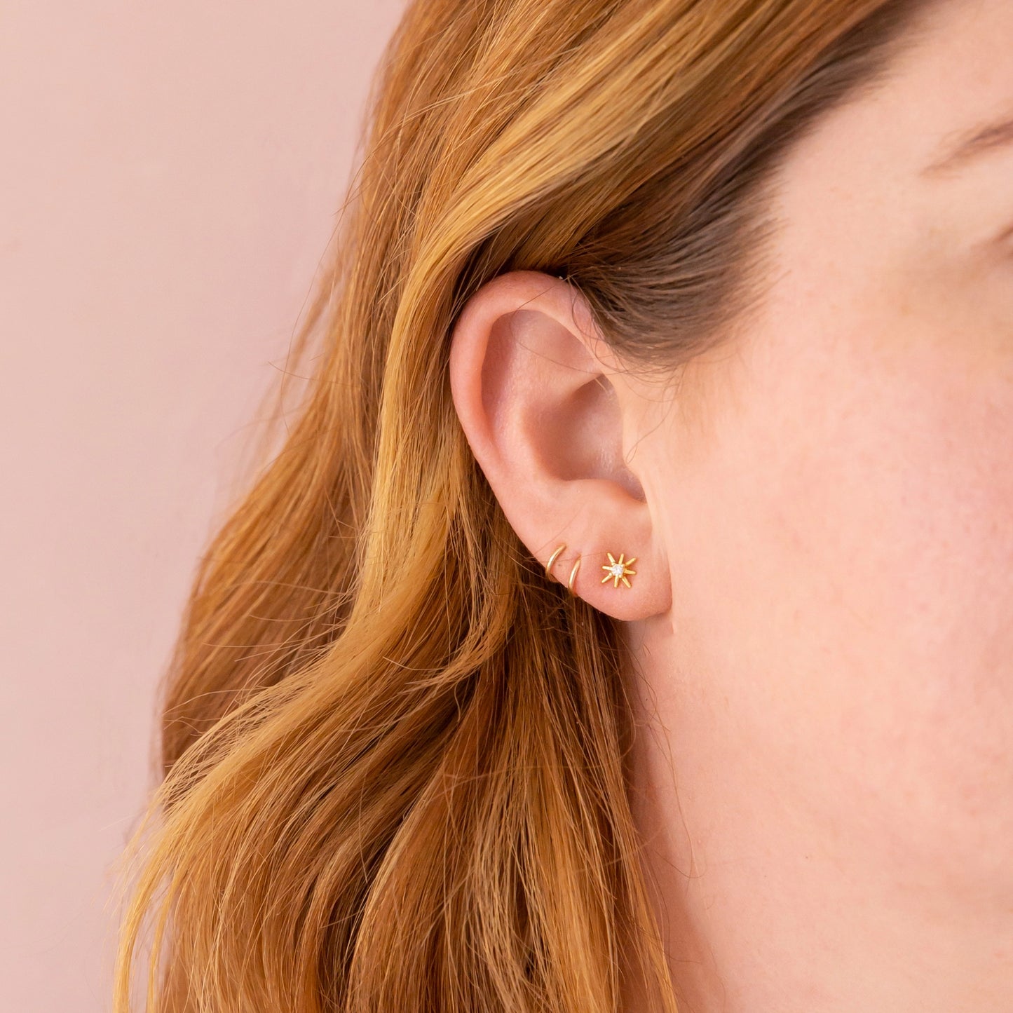 On a peachy background is a model wearing the dainty star earrings along with an existing earring stack. 