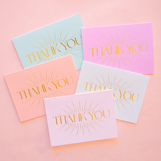 On an ivory background is five different colored thank you cards with gold foiled letters in the center that read, "Thank You". The colors come in fuchsia, mint green, light pink, white and salmon. 