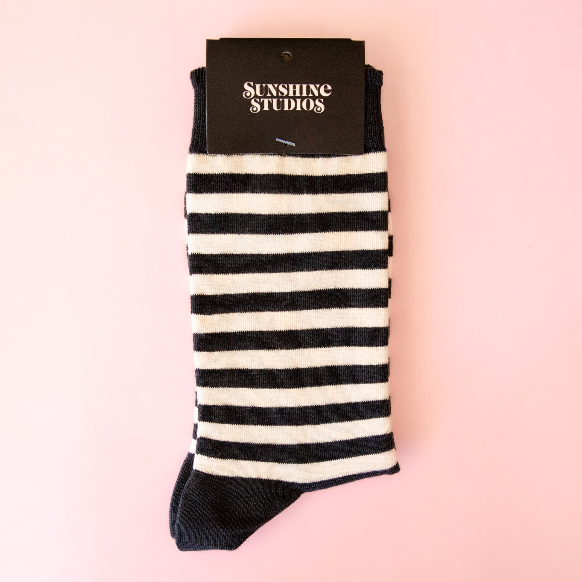 A pair of black and white striped socks on a pink background with packaging that reads, "sunshine studios".
