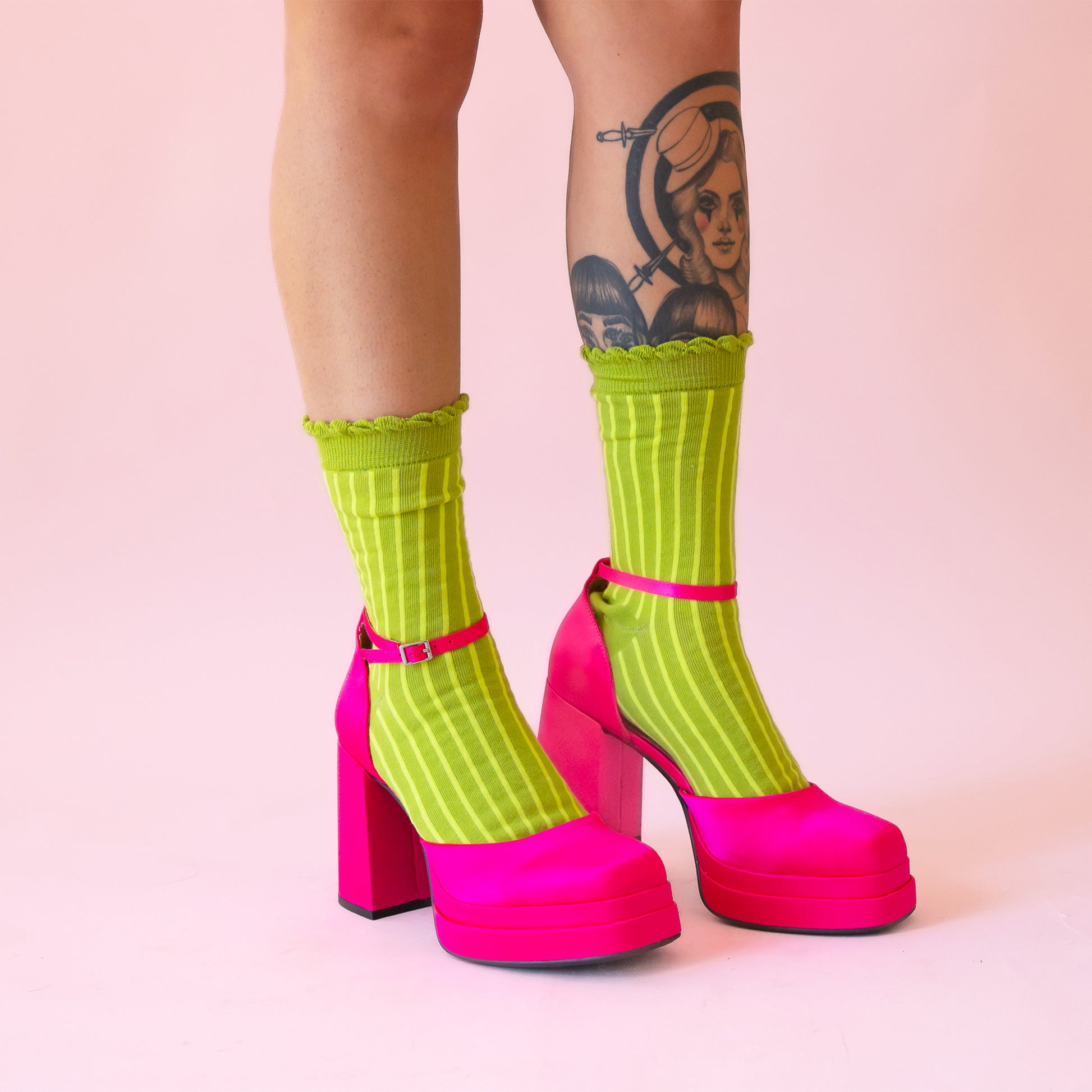 A pair of lime green crew socks with a lighter green stripe and a ruffle edge at the top. Worn on a model with hot pink satin closed toe high heels. 