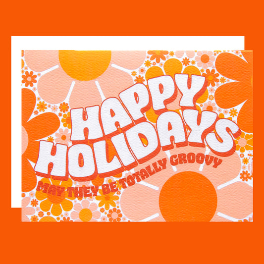 Card filled with a funky 70’s design made up of orange and peach daisies. On top of the design is white block lettering with a red shadow that reads ‘Happy Holidays.’ Under that in red block text it reads ‘may they be totally groovy.’ This card is accompanied by a white envelope.