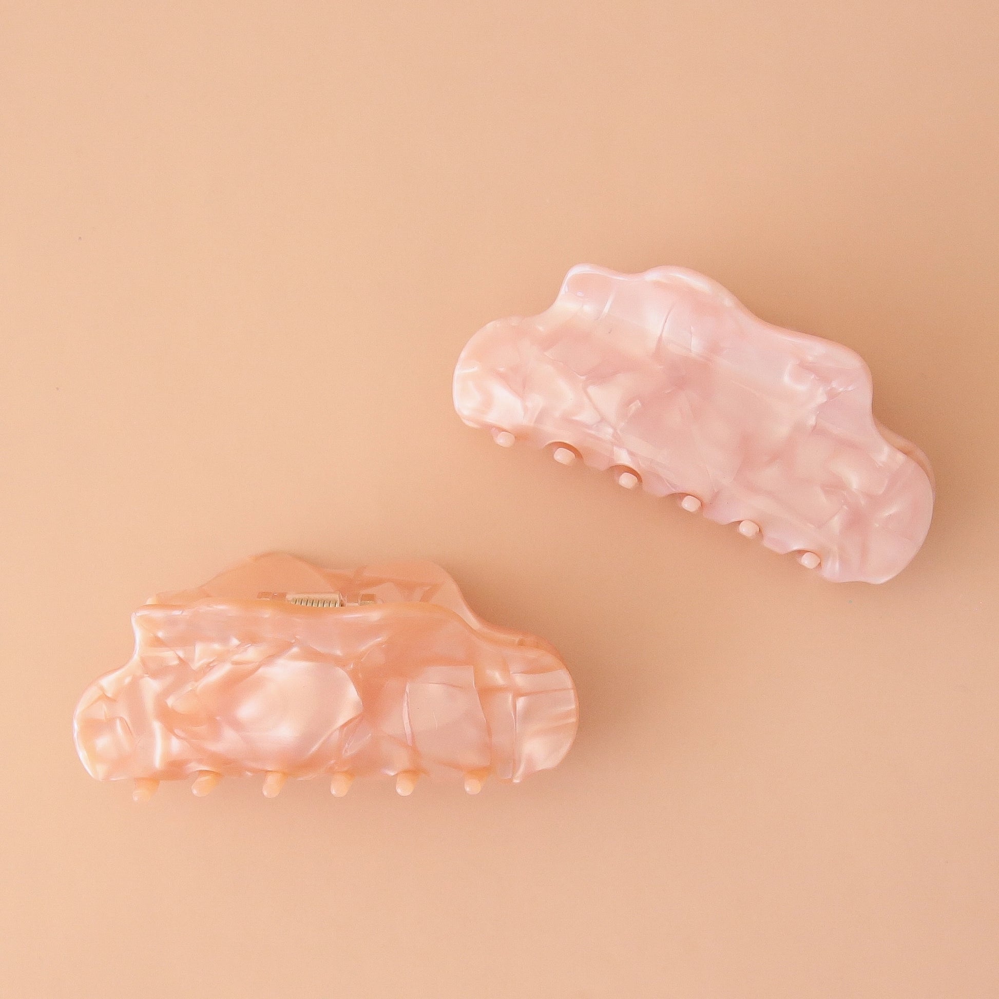 On a peachy background is two shell textured claw clips with a wavy edge detail in a peachy shade and a light pink shade.