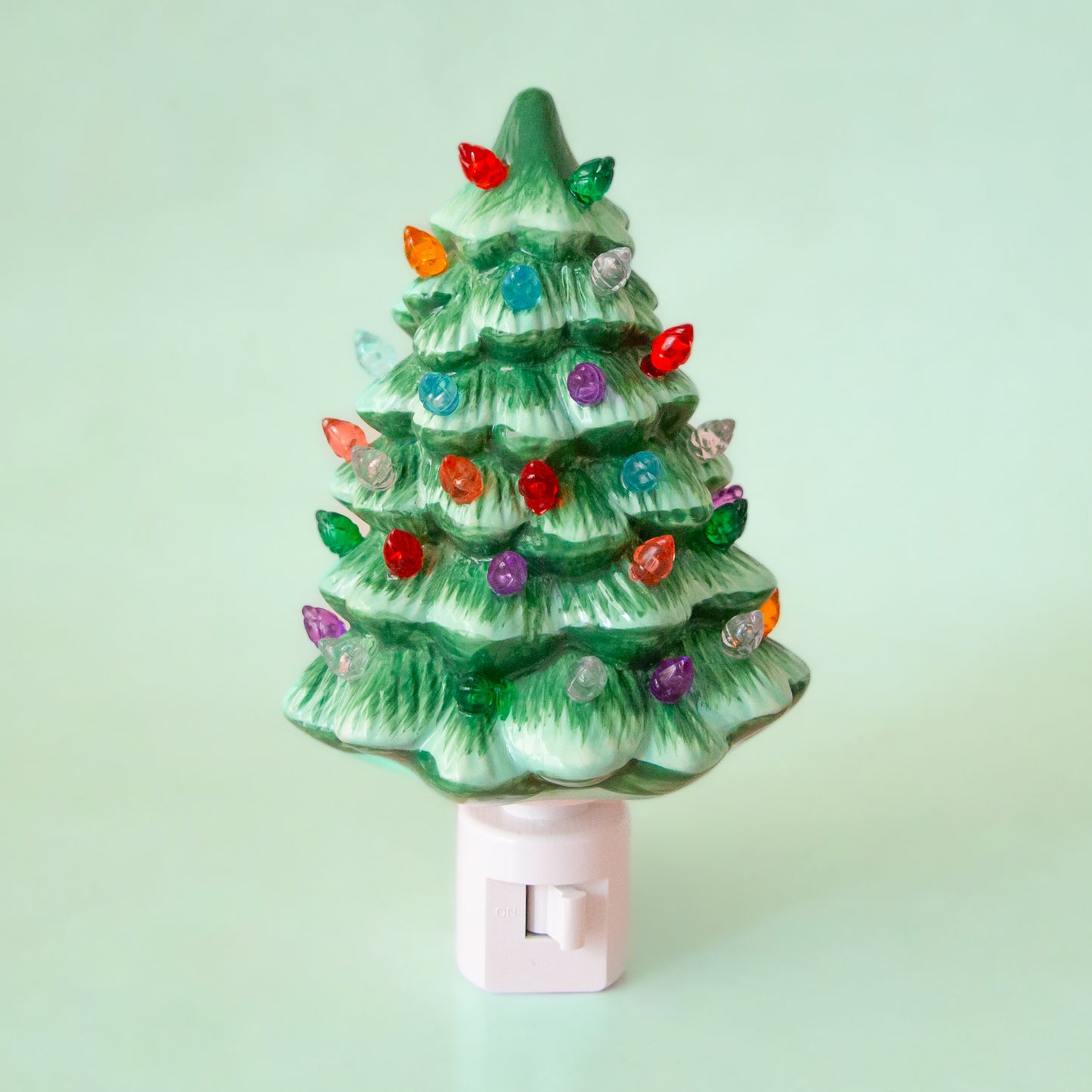 A ceramic Christmas Tree shaped night light sits in middle of the page with bright colorful lights.  It has a white plug at the bottom.  The tree is a green color.