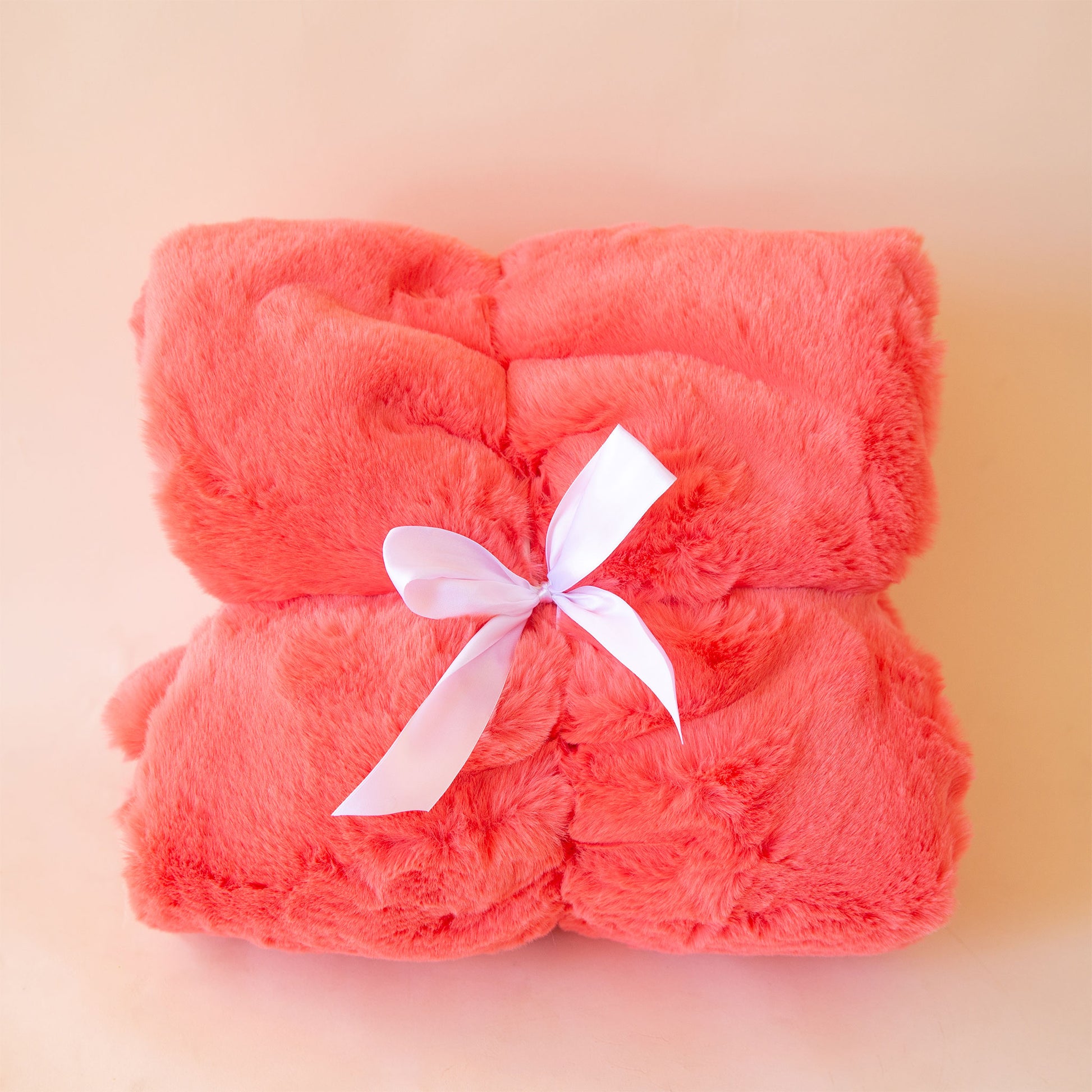 On a peach background is a faux fur reddish pink blanket folded and tied with a white satin ribbon.