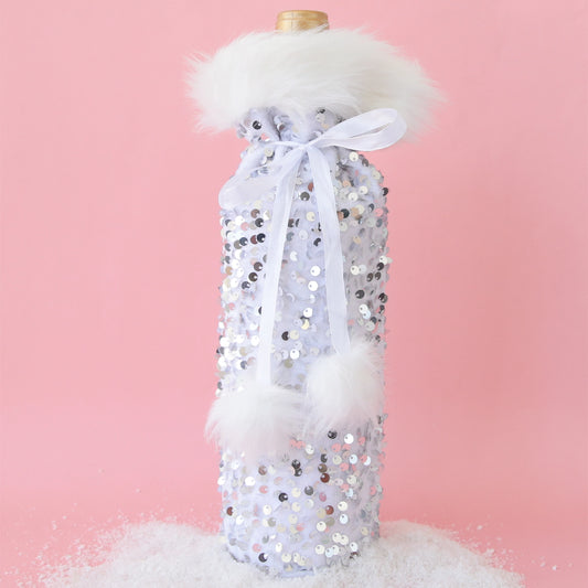 On a pink background is a silver sequin wine gift bag with white faux fur around the opening and a white pom pom bow around the top.