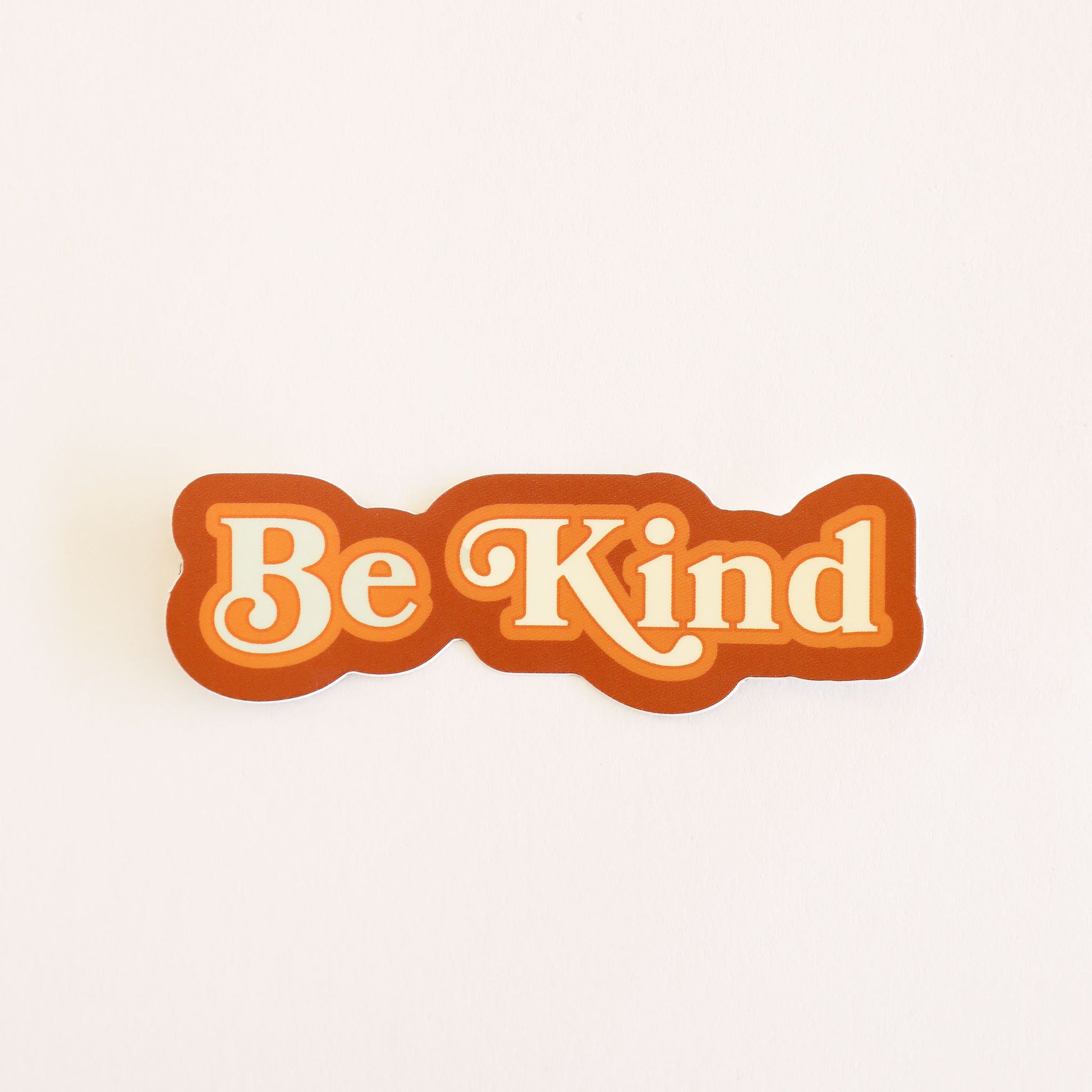 Sticker reading 'Be Kind' in white lettering, outlined in tangerine orange and rusty red. 