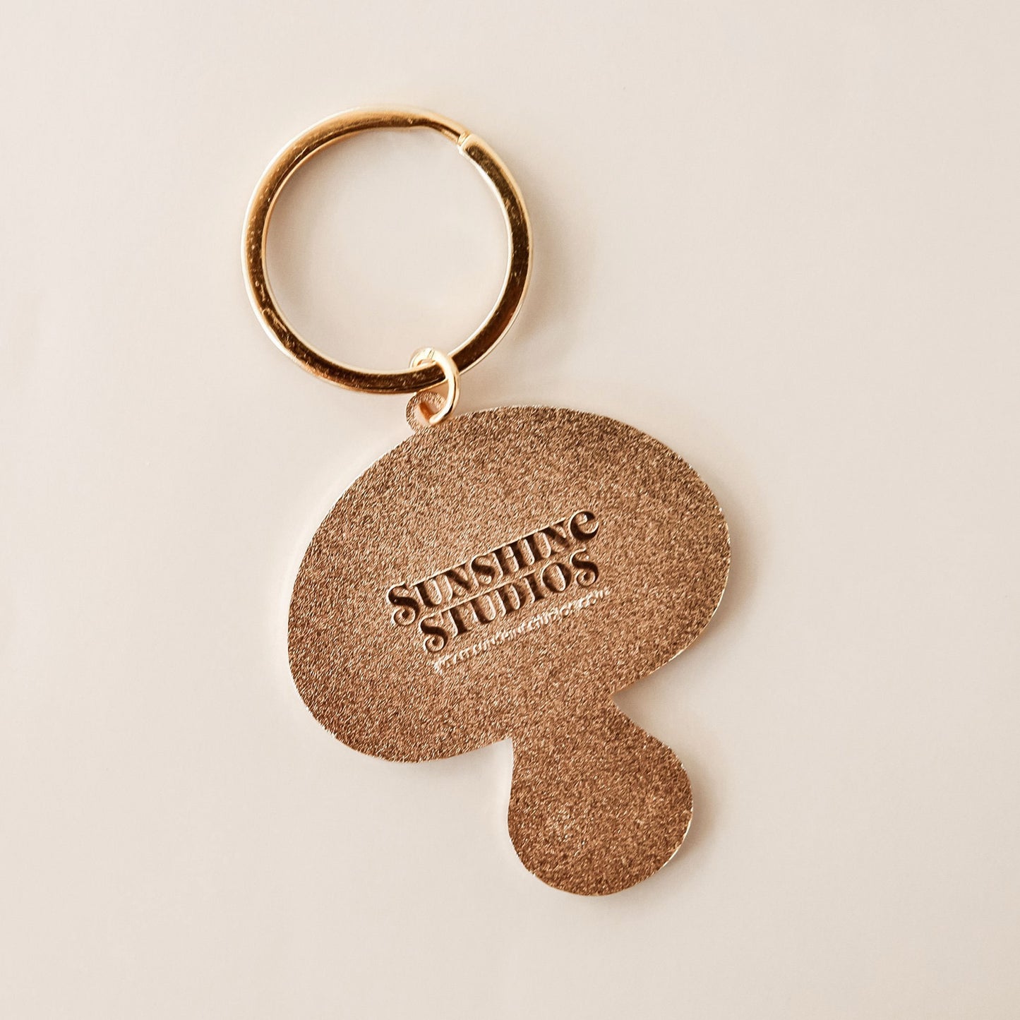 Backside of mushroom keychain is golden and textured, reading 'sunshine studios' in raised lettering.