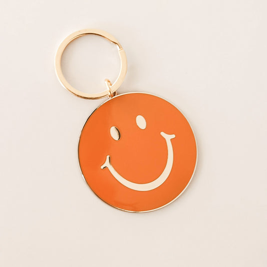 Orange circle keychain with gold smiley face design. The keychain is complete with a golden key chain ring.