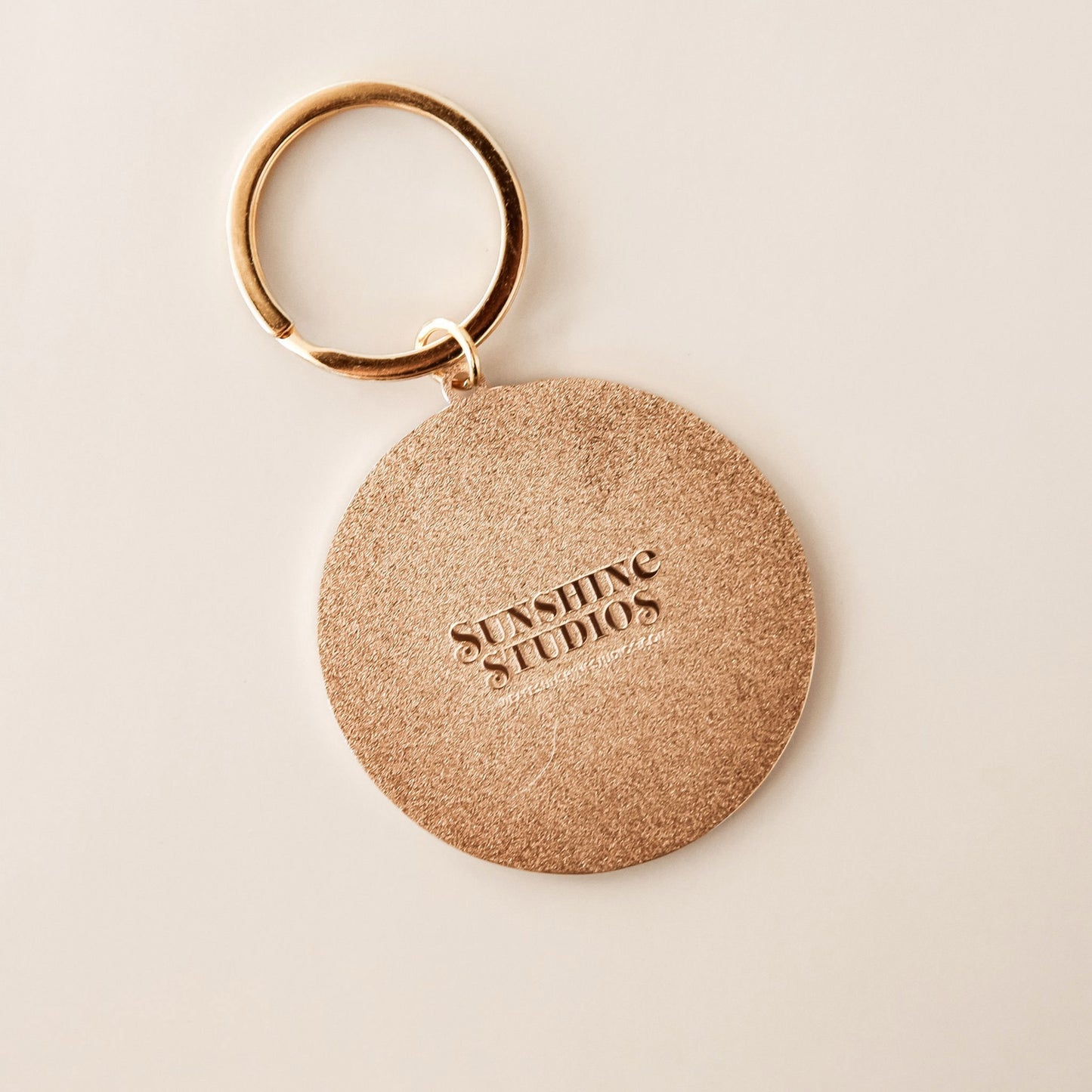 Textured backside of round keychain reading 'sunshine studios' in raised lettering. The keychain is complete with a golden keychain loop. 