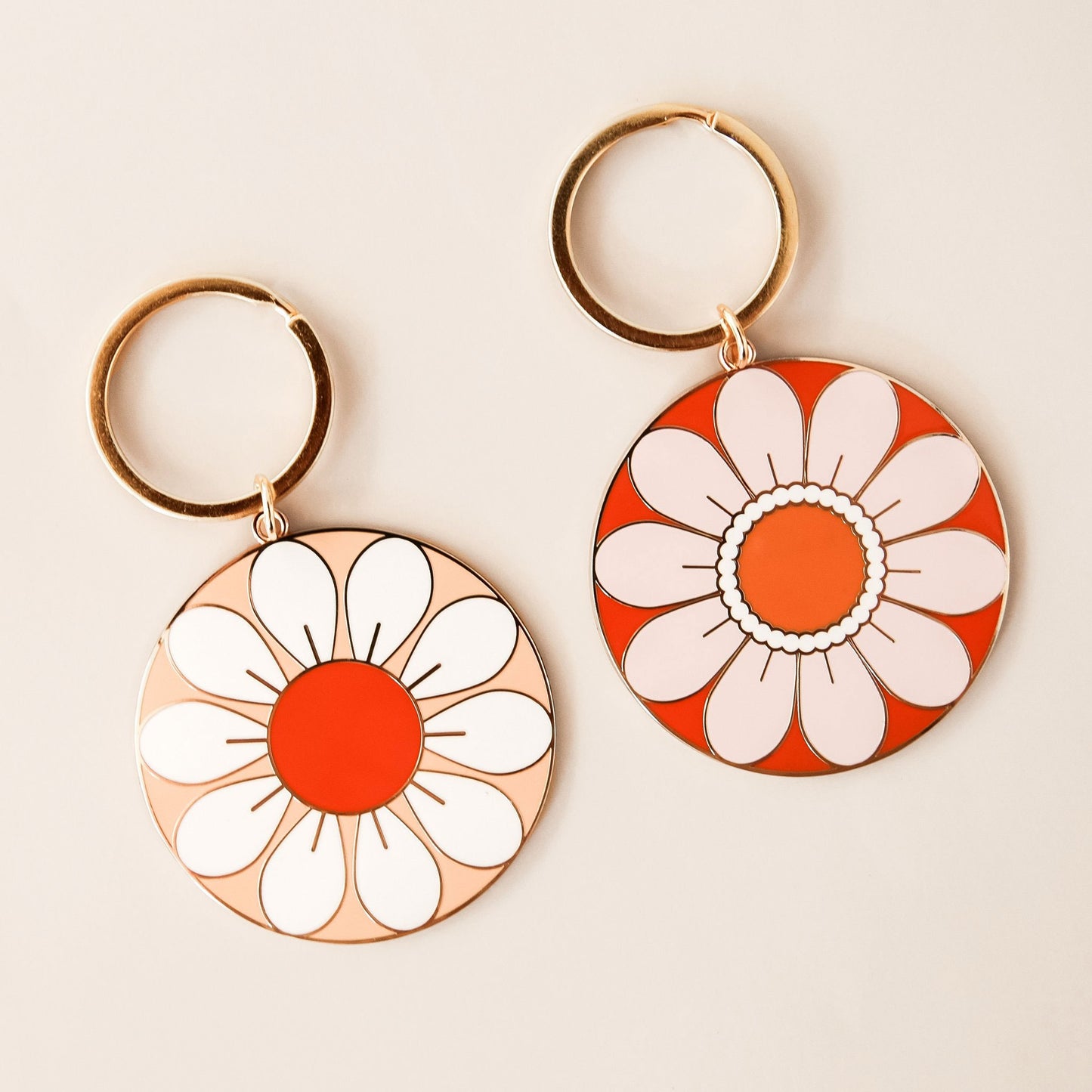 Two circular flower key chains both complete with golden key chain hoops. To the left is a flower with white petals and red center and to the right is a flower with light beige petals and orange center.