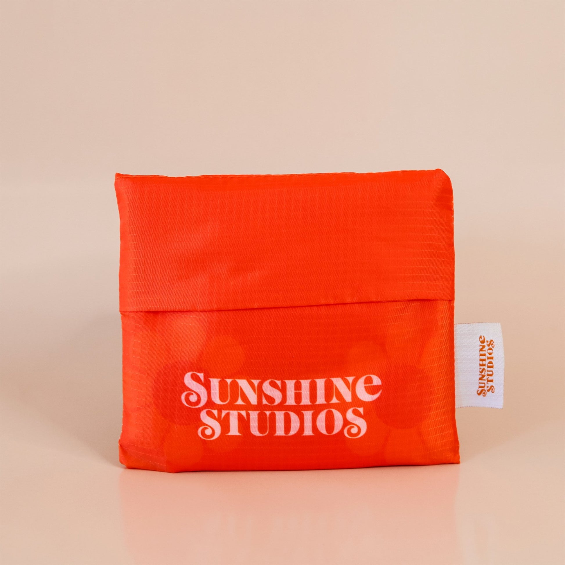 Bright orange reusable bag folded into a square. At the bottom there is peach text that reads ’sunshine studios.’ To the right is a white tag with bright orange text that reads ’sunshine studios.’