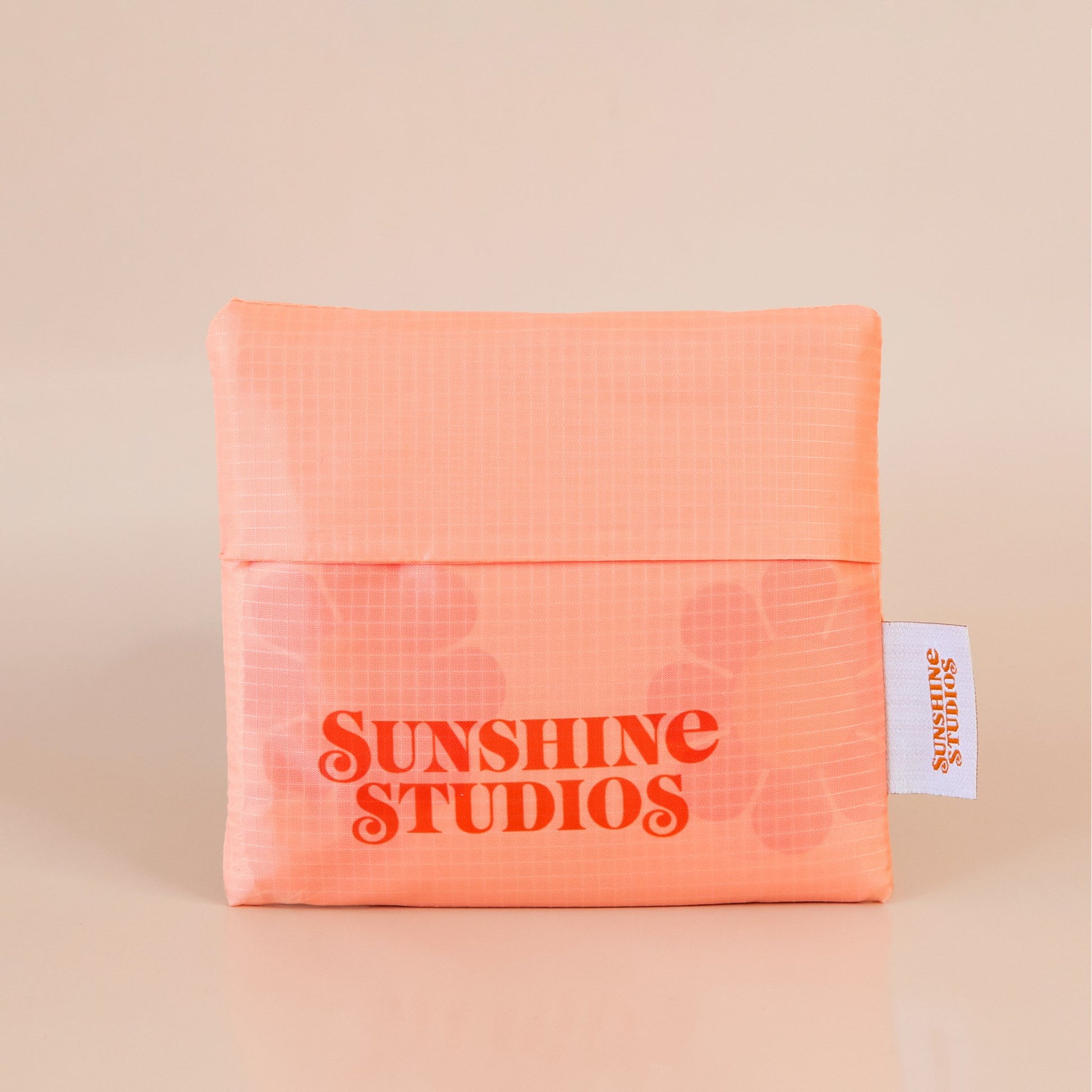 Peach reusable bag folded into a square. At the bottom there is red-orange text that reads ’sunshine studios.’ To the right is a white tag with bright orange text that reads ’sunshine studios.’