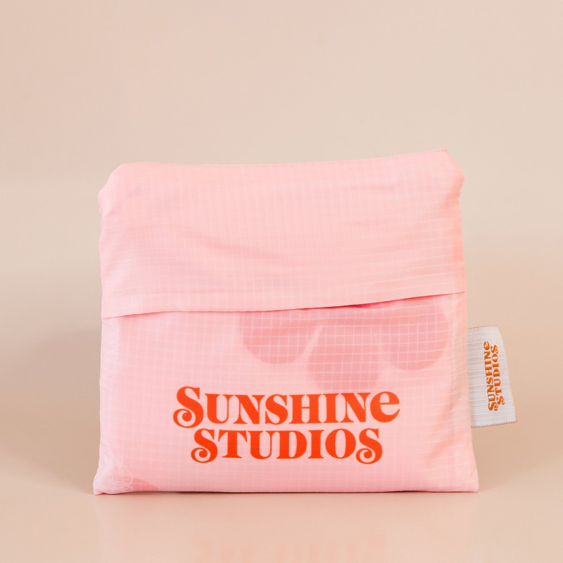 Soft pink reusable bag folded into a square. At the bottom there is peach text that reads ’sunshine studios.’ To the right is a white tag with bright orange text that reads ’sunshine studios.’ 