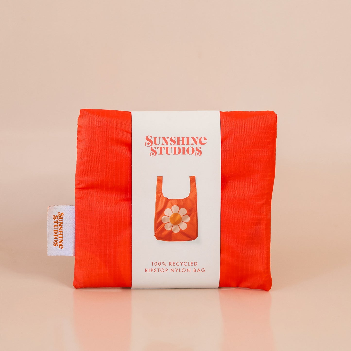 Red-orange reusable bag folded tightly into a square. The bag is wrapped in a white band that reads ’sunshine studios'. Under the text is a picture of the reusable bag. On the left side of the bag is a white tag with bright orange text that reads ’sunshine studios.' 