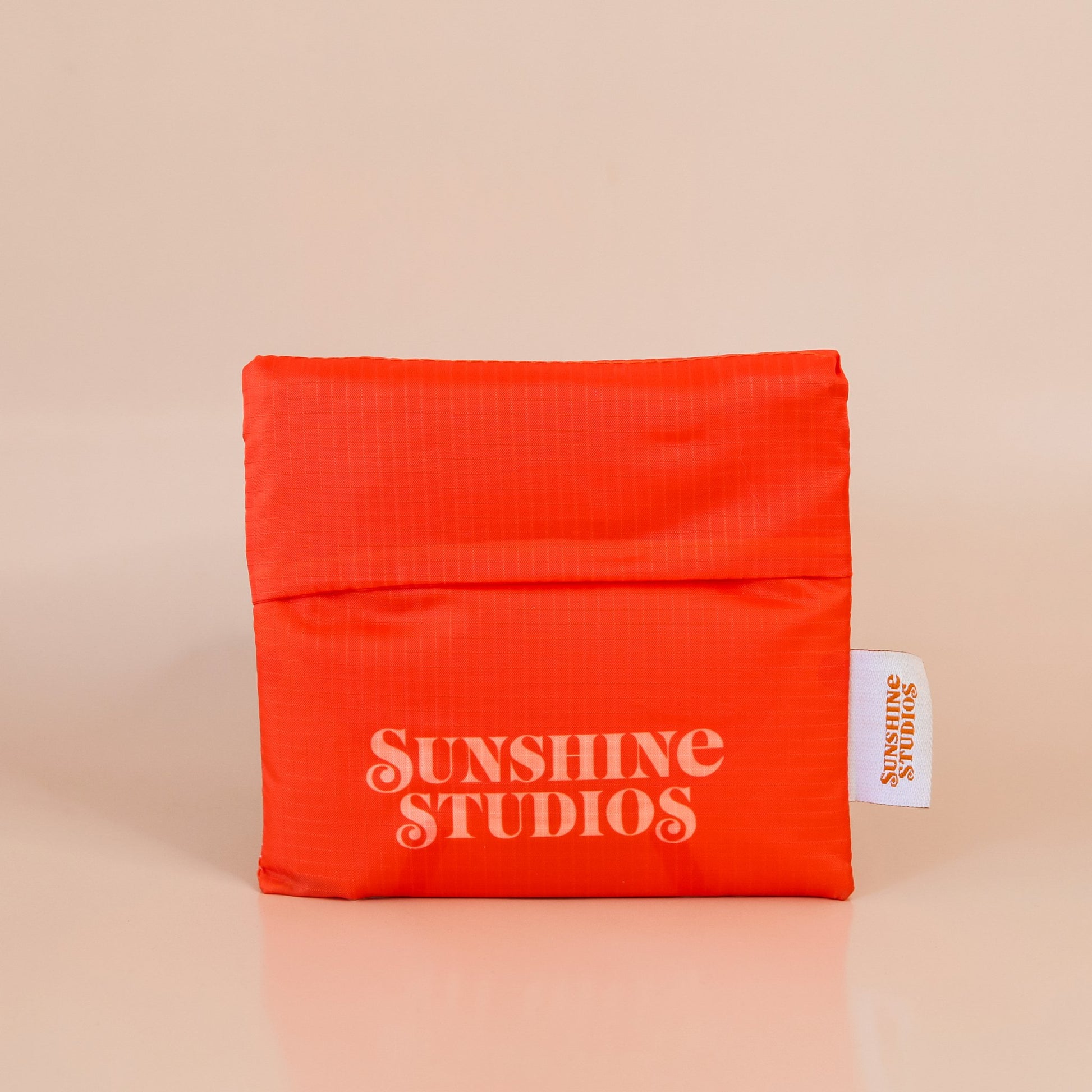 Red-orange nylon bag folded up into a compact square. The bag reads 'sunshine studios' in off white lettering and has a small white tag that reads 'sunshine studios' in orange lettering. 