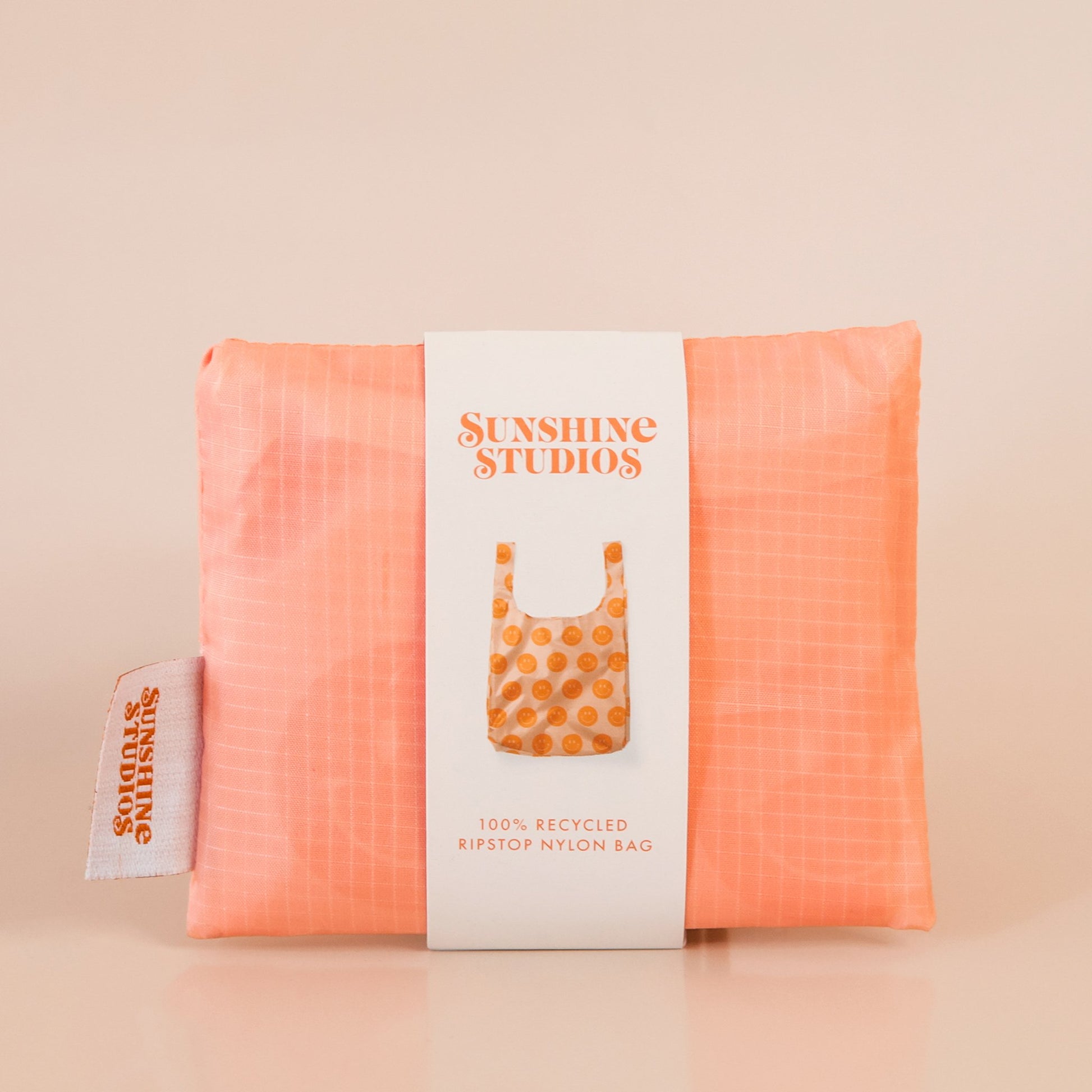 Folded up peach reusable bag folded into a small square and wrapped with white packaging. The packaging reads 'sunshine studios'. The label has a photo of the bag unfolded and reads '100% recycled ripstop nylon bag'