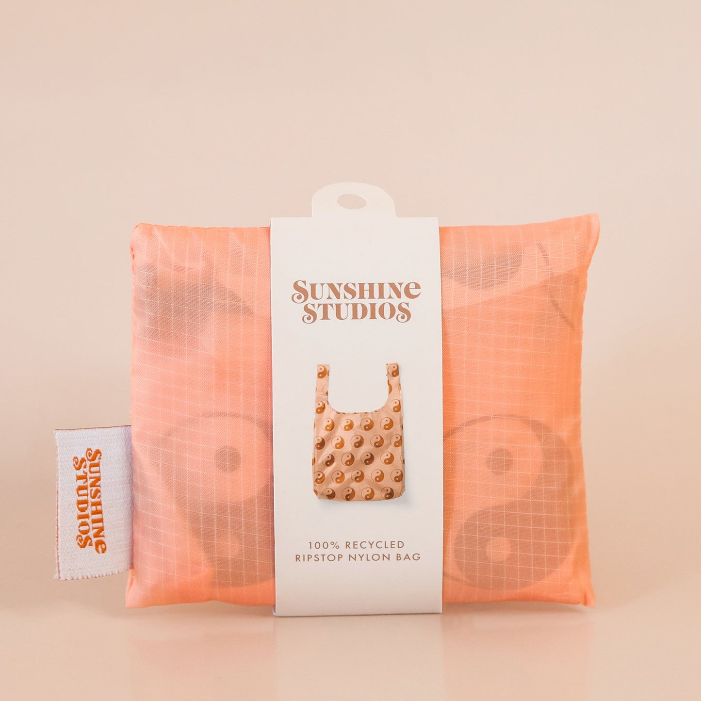 Peach reusable bag folded tightly into a square. The bag is wrapped in a white band that reads ’sunshine studios'. Under the text is a picture of the reusable bag. On the left side of the bag is a white tag with bright orange text that reads ’sunshine studios.'