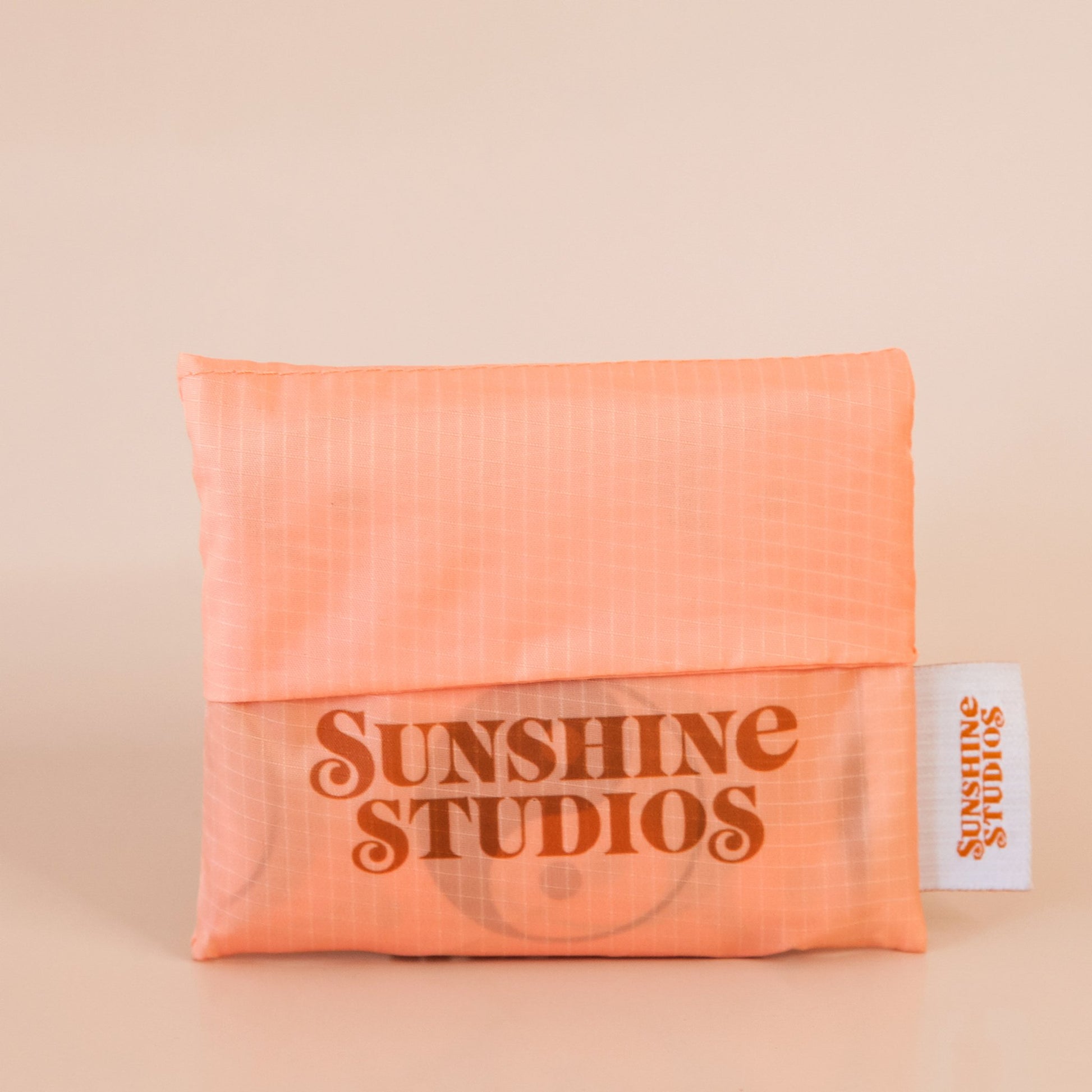 Peach reusable bag folded into a square. At the bottom there is red-orange text that reads ’sunshine studios.’ To the right is a white tag with bright orange text that reads ’sunshine studios.’