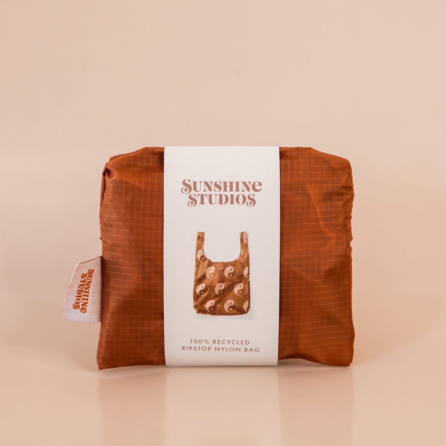 Chocolate brown reusable bag folded tightly into a square. The bag is wrapped in a white band that reads ’sunshine studios'. Under the text is a picture of the reusable bag. On the left side of the bag is a white tag with bright orange text that reads ’sunshine studios.'