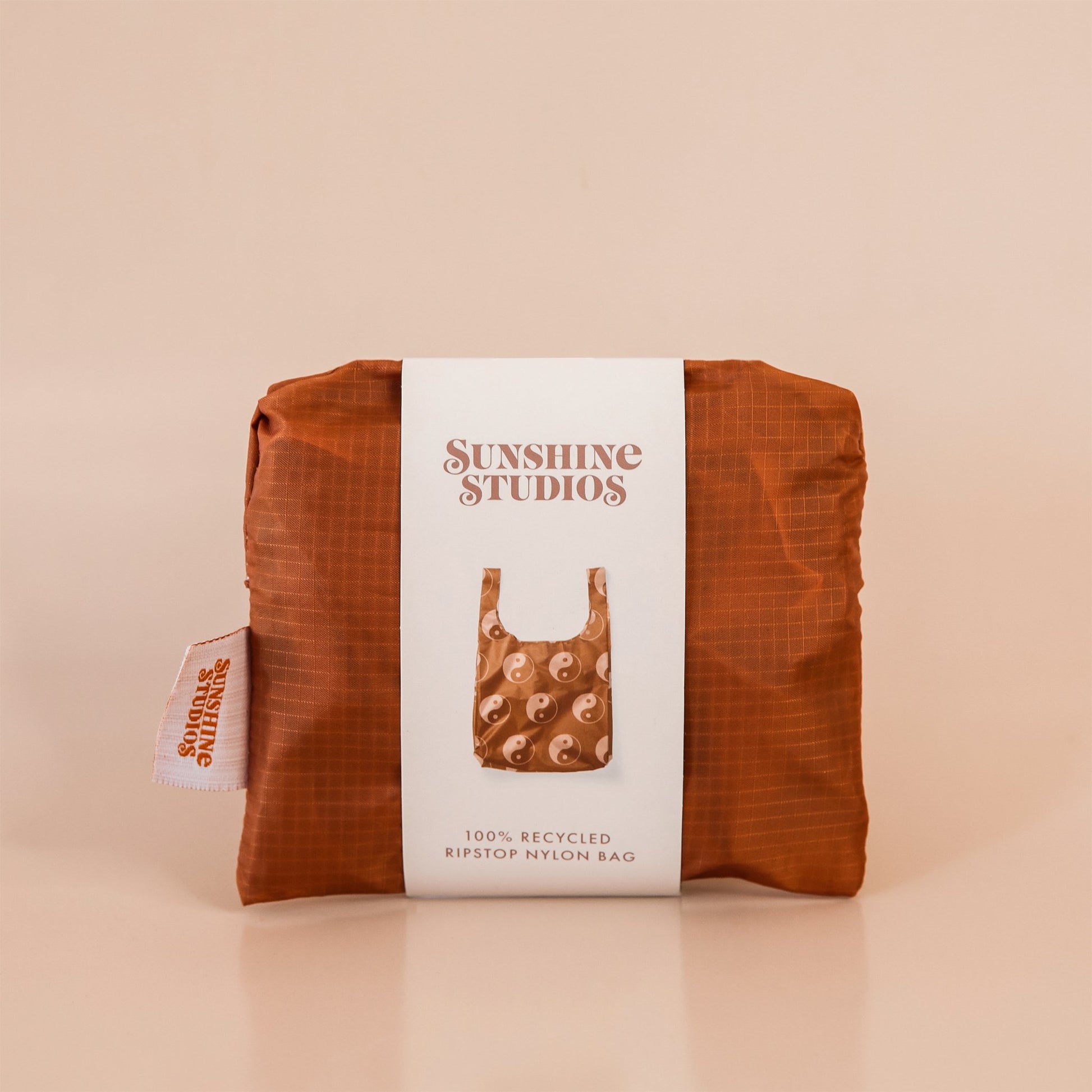 Chocolate brown reusable bag folded tightly into a square. The bag is wrapped in a white band that reads ’sunshine studios'. Under the text is a picture of the reusable bag. On the left side of the bag is a white tag with bright orange text that reads ’sunshine studios.'
