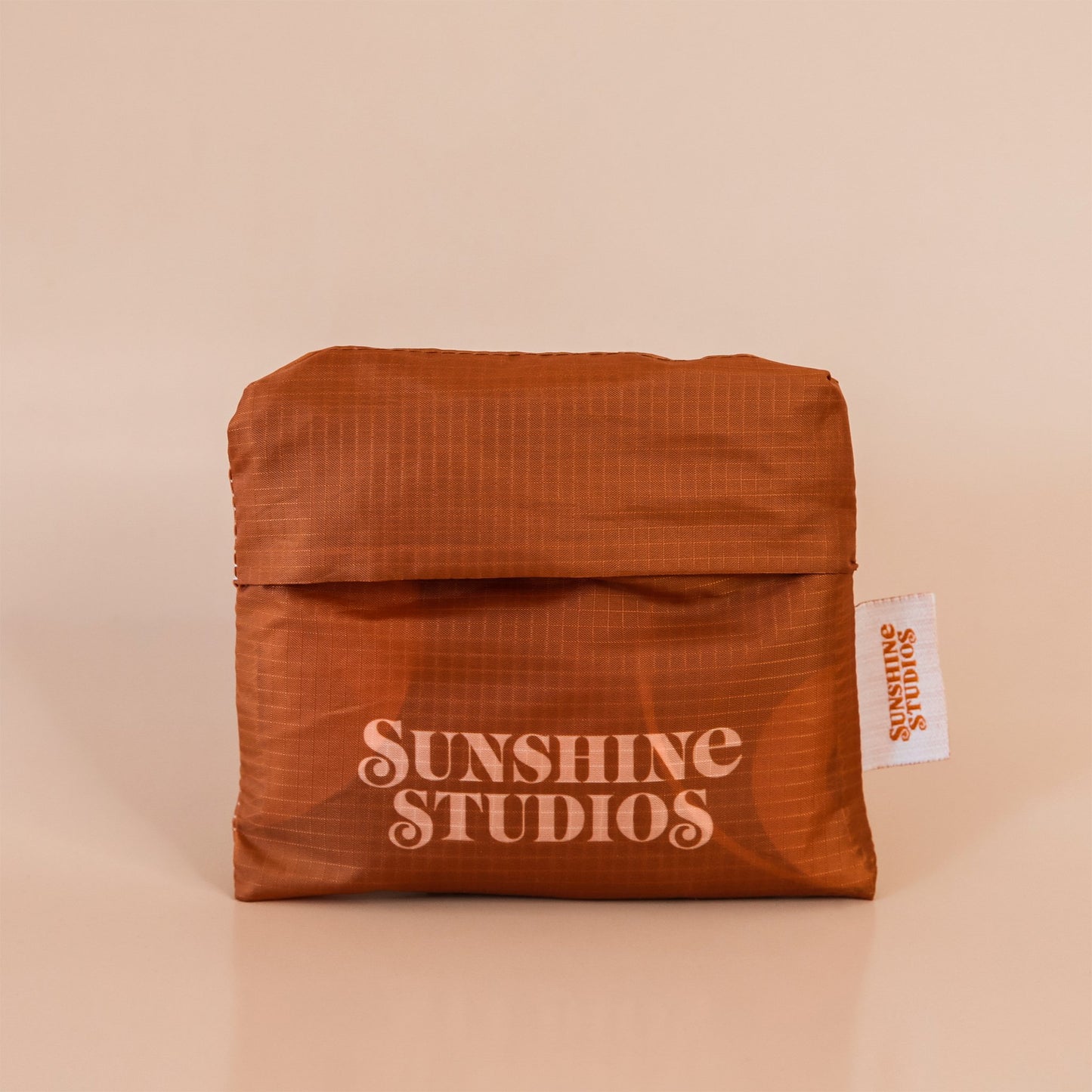 Brown reusable bag folded into a square. At the bottom there is peach text that reads ’sunshine studios.’ To the right is a white tag with bright orange text that reads ’sunshine studios.’