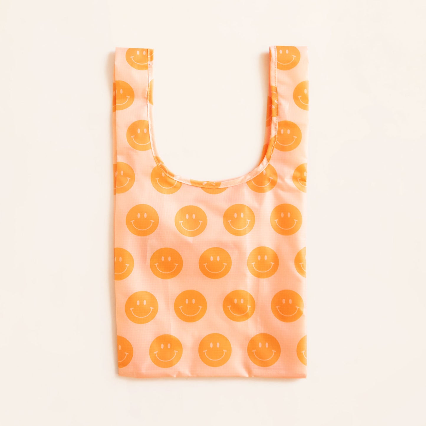 Peach reusable covered in a print of orange smiley facies. The bag is positioned flat on a table and has a 'U' shape between two handles.