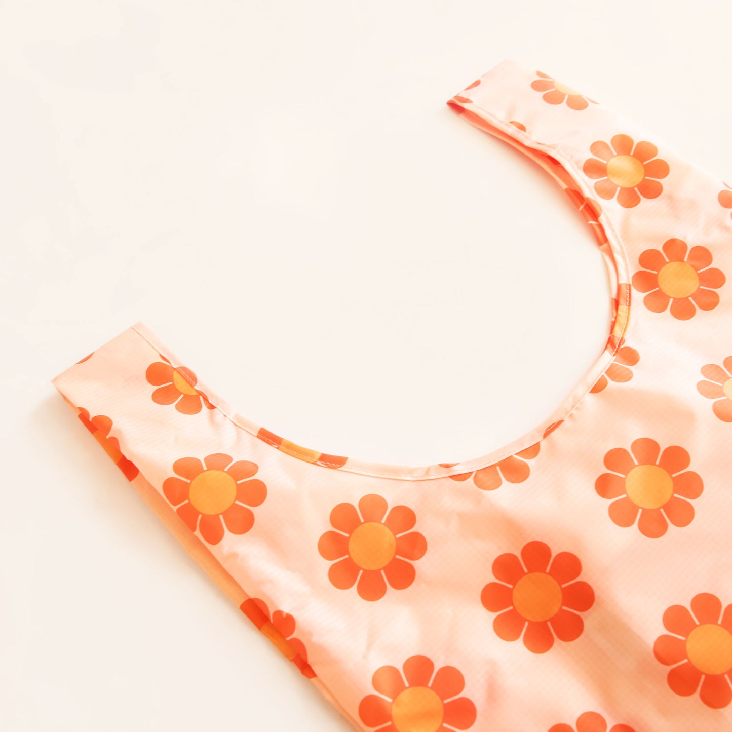Peach reusable bag filled with a pattern of flowers with red-orange petals and tangerine centers. The bag is positioned flat on a table and has a 'U' shape between two handles. 