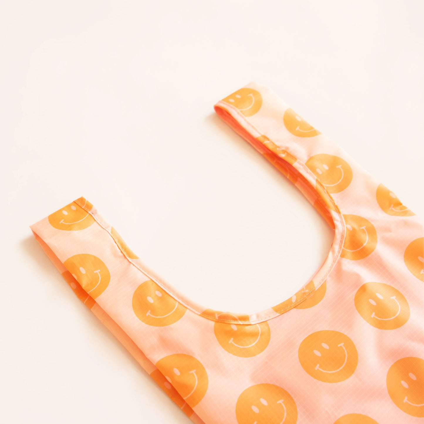 Zoomed view of peach nylon reusable bag. The bag is covered in a print of orange smiley faces. 