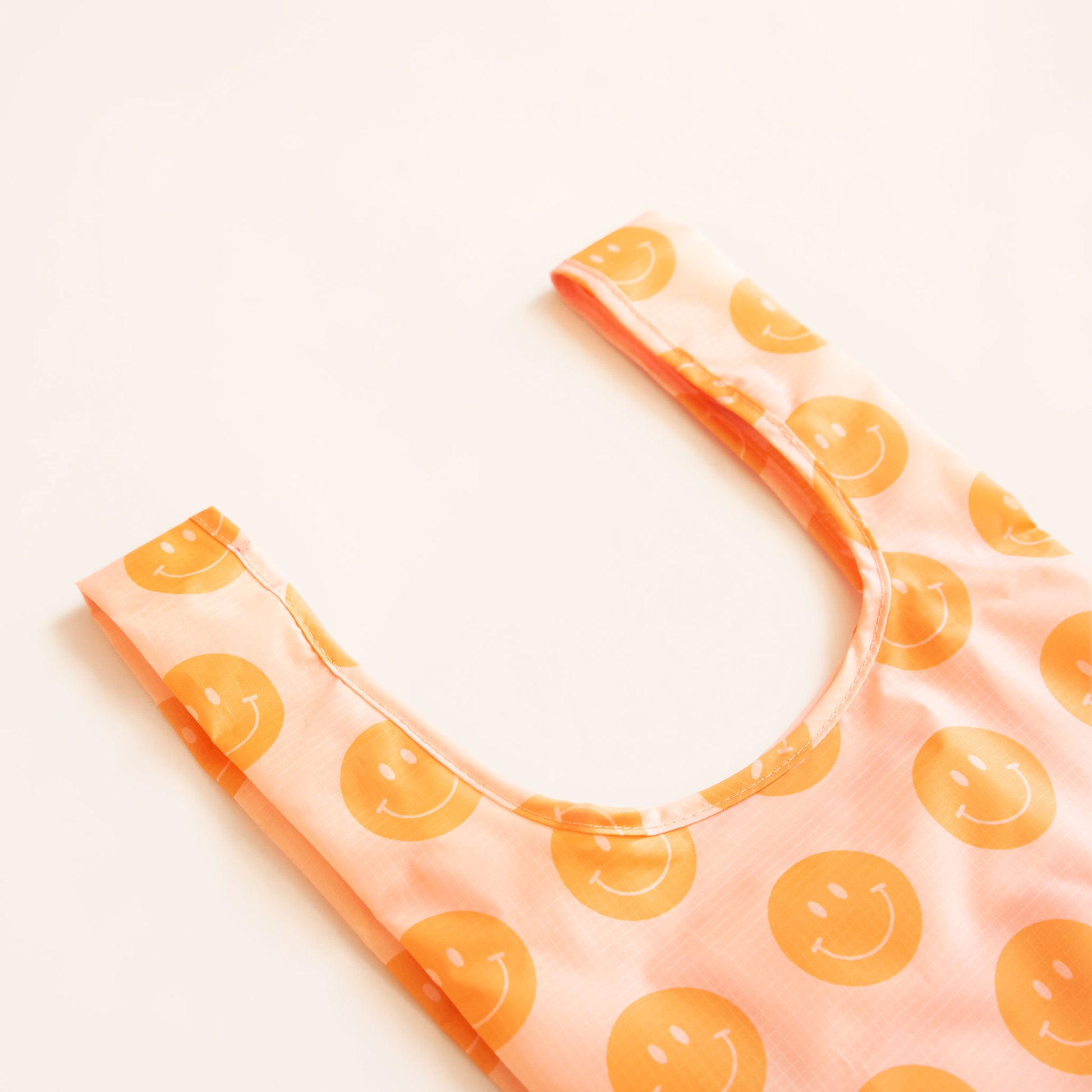 Zoomed view of peach nylon reusable bag. The bag is covered in a print of orange smiley faces. 