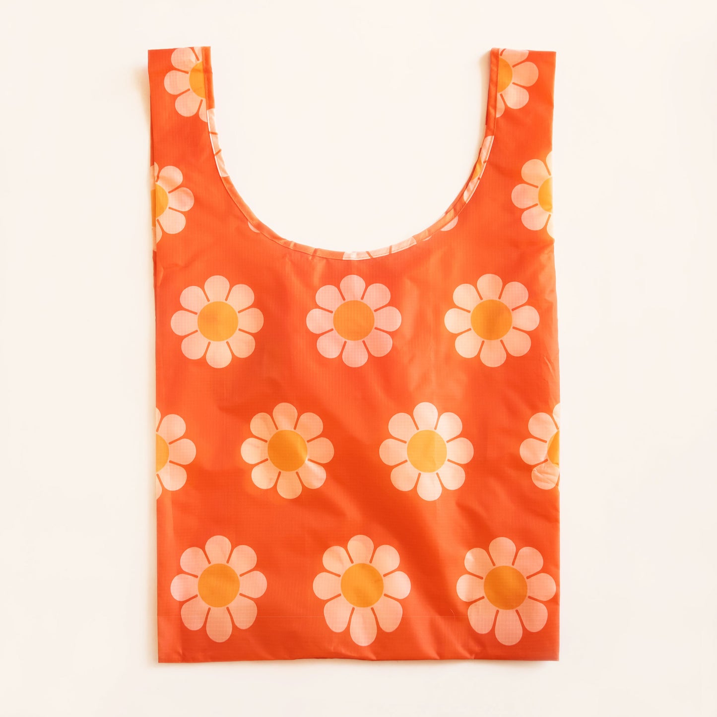 Red-orange reusable covered in a print of simple flowers with white petals and orange centers. The bag is positioned flat on a table and has a 'U' shape between two handles. 