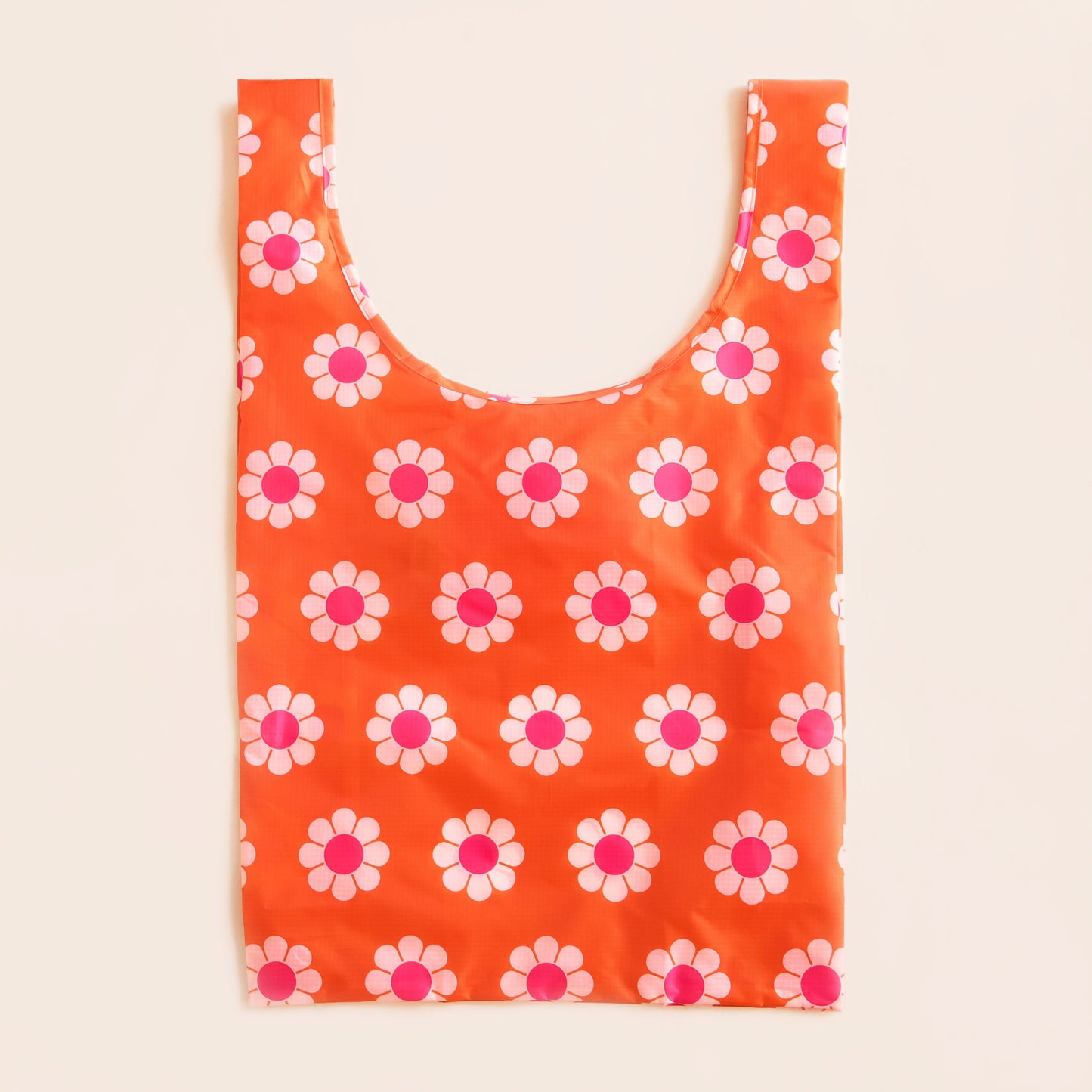 Red-orange reusable bag covered in a print of simple flowers with white petals and pink centers. The bag is positioned flat on a table and has a 'U' shape between two handles. 