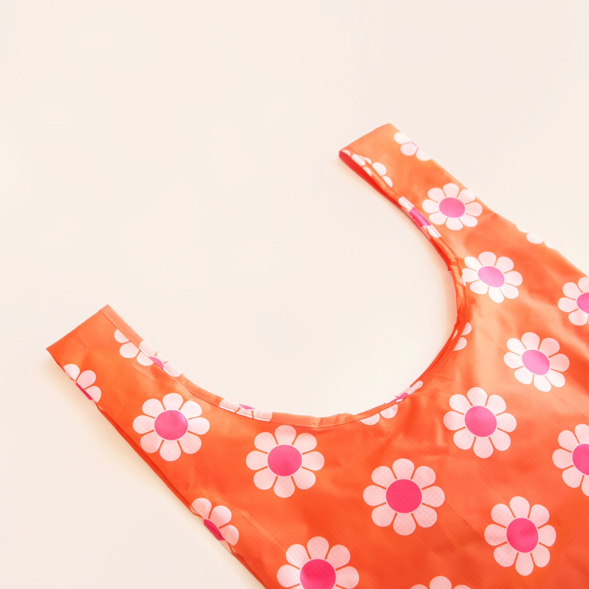 Red-orange reusable bag covered in a print of simple flowers with white petals and pink centers. The bag is positioned flat on a table and has a 'U' shape between two handles.