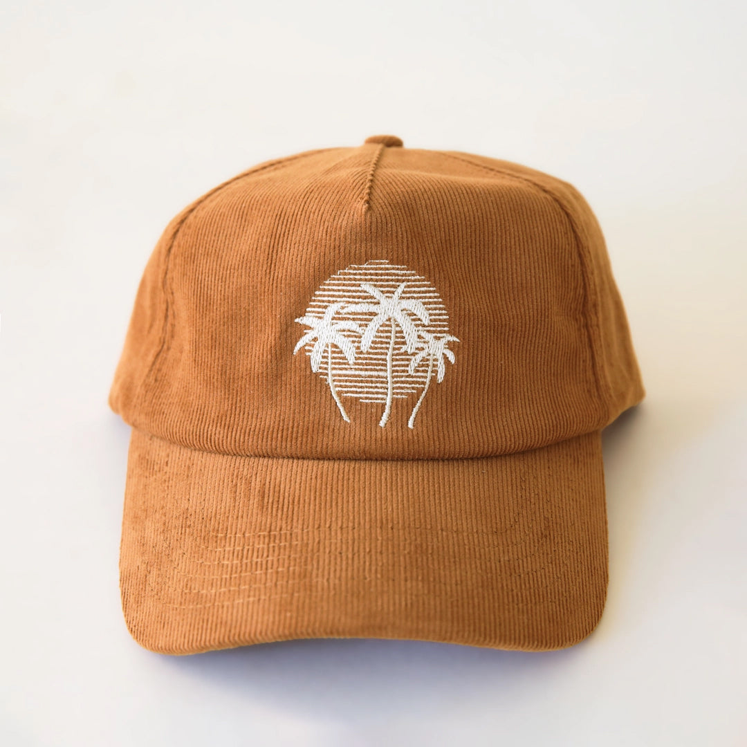 On a cream background is a rust colored corduroy snapback hat with a curved bill and a circle graphic with three white palm trees.