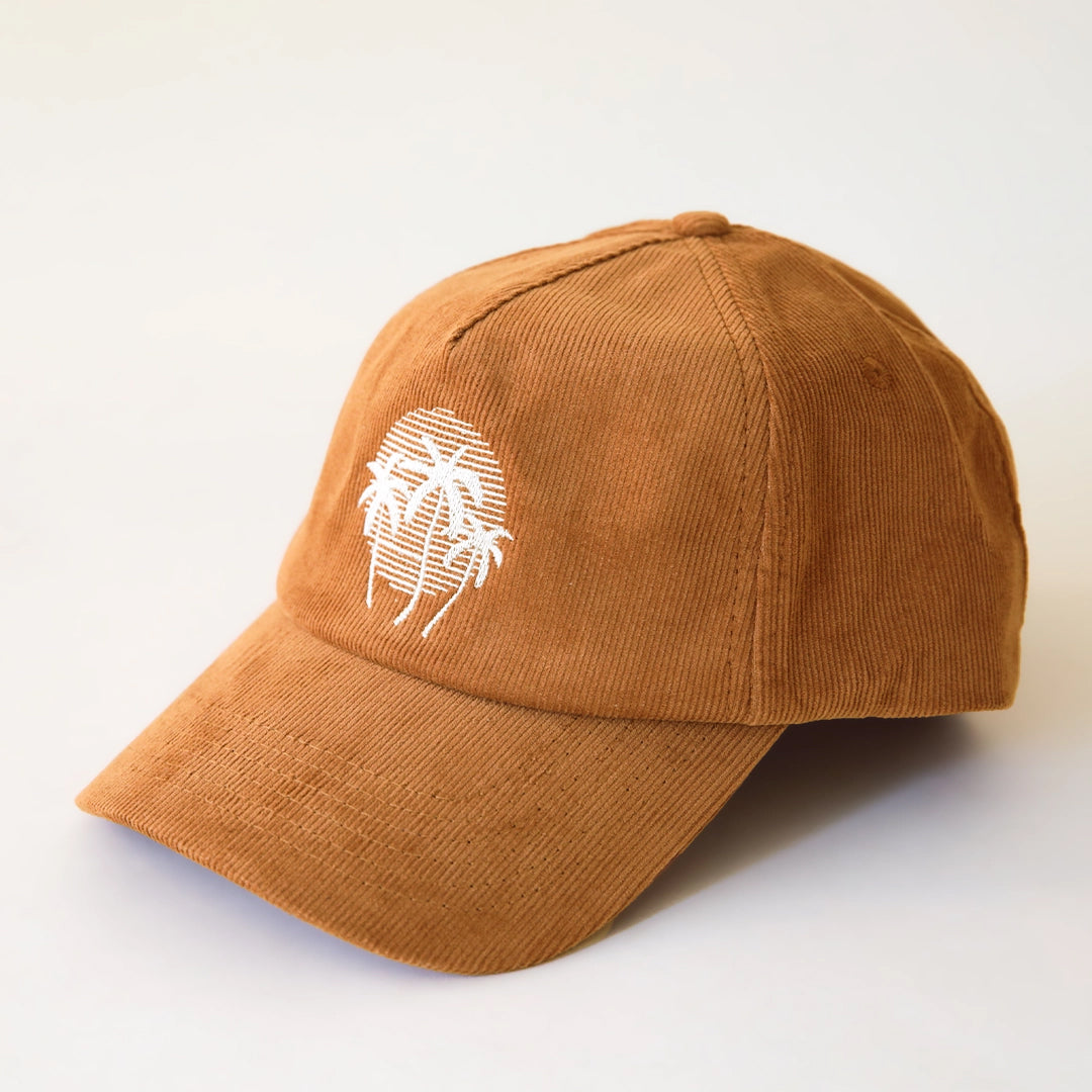 On a cream background is a rust colored corduroy snapback hat with a curved bill and a circle graphic with three white palm trees.