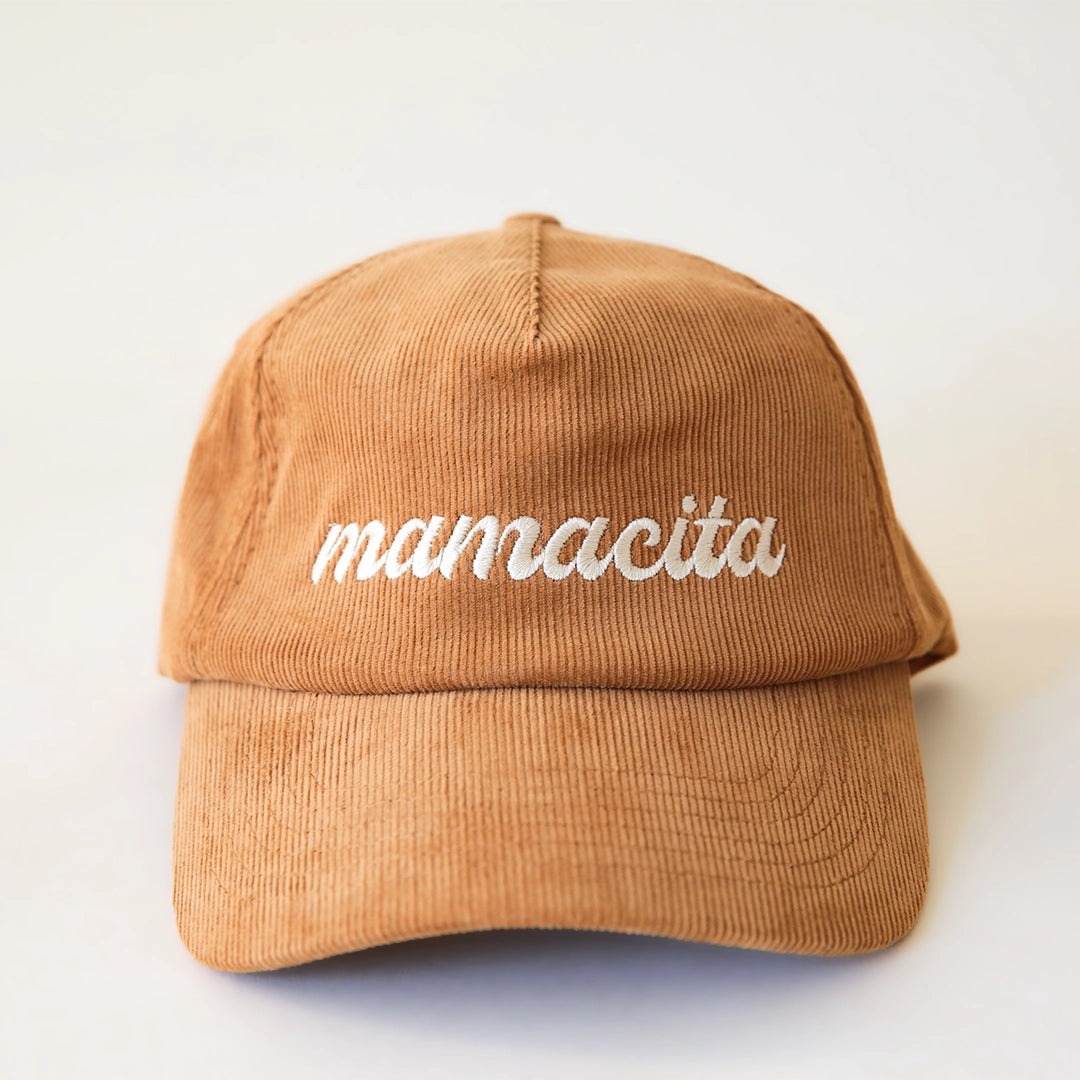 On a cream background is a  burnt orange corduroy snapback hat with white cursive text that reads, "mamacita".