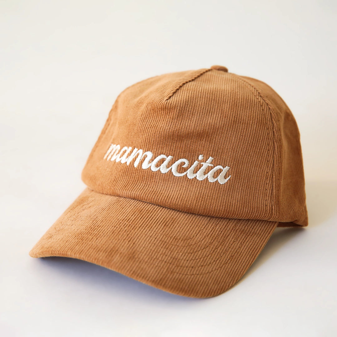 On a cream background is a burnt orange corduroy snapback hat with white cursive text that reads, "mamacita".