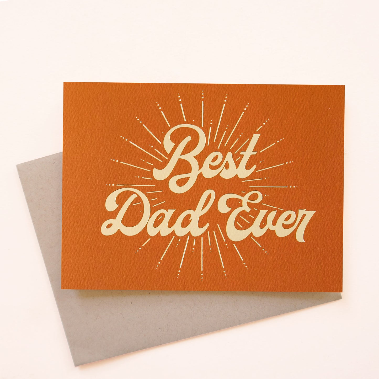 Burnt orange greeting card reading 'Best Dad Ever' in light cursive lettering. A sun burst design accents the text. The card is accompanied by a Kraft brown envelope. 